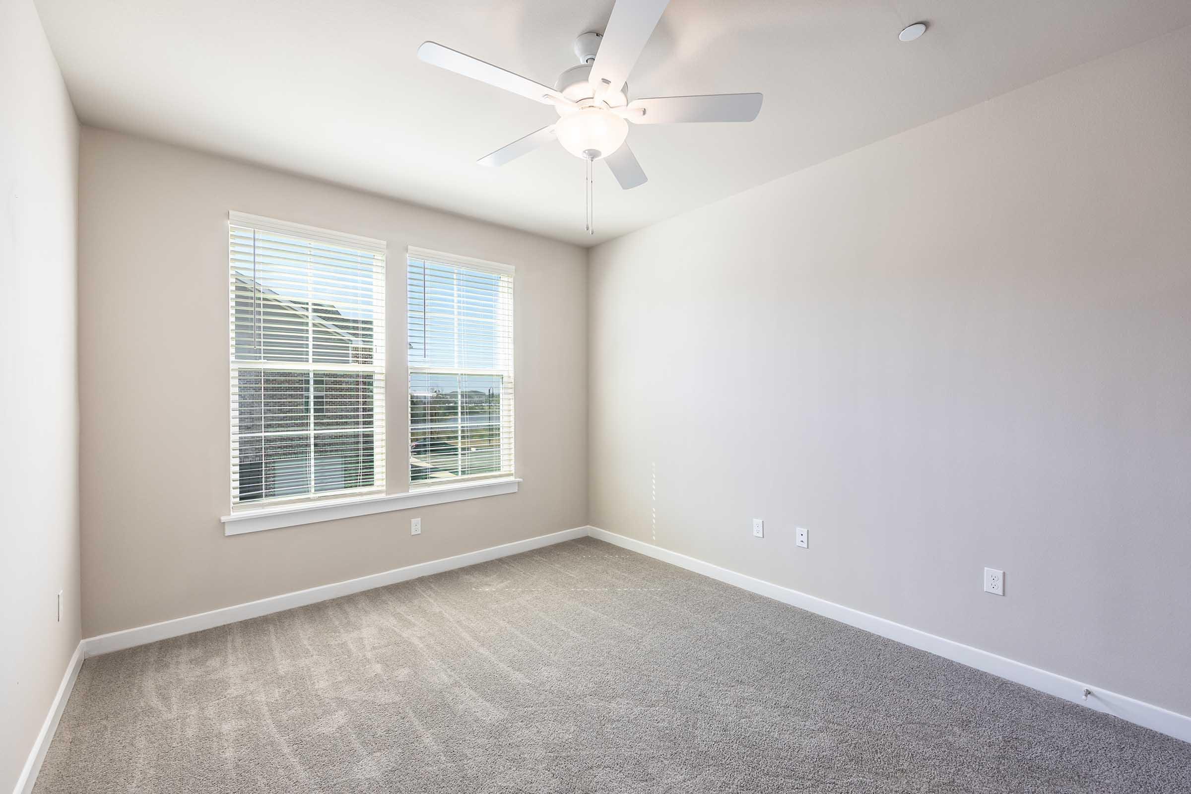A vacant room featuring light-colored walls, a ceiling fan, and large windows with white blinds. The floor is carpeted in a neutral tone, and there are no furnishings, creating a bright and spacious ambiance.