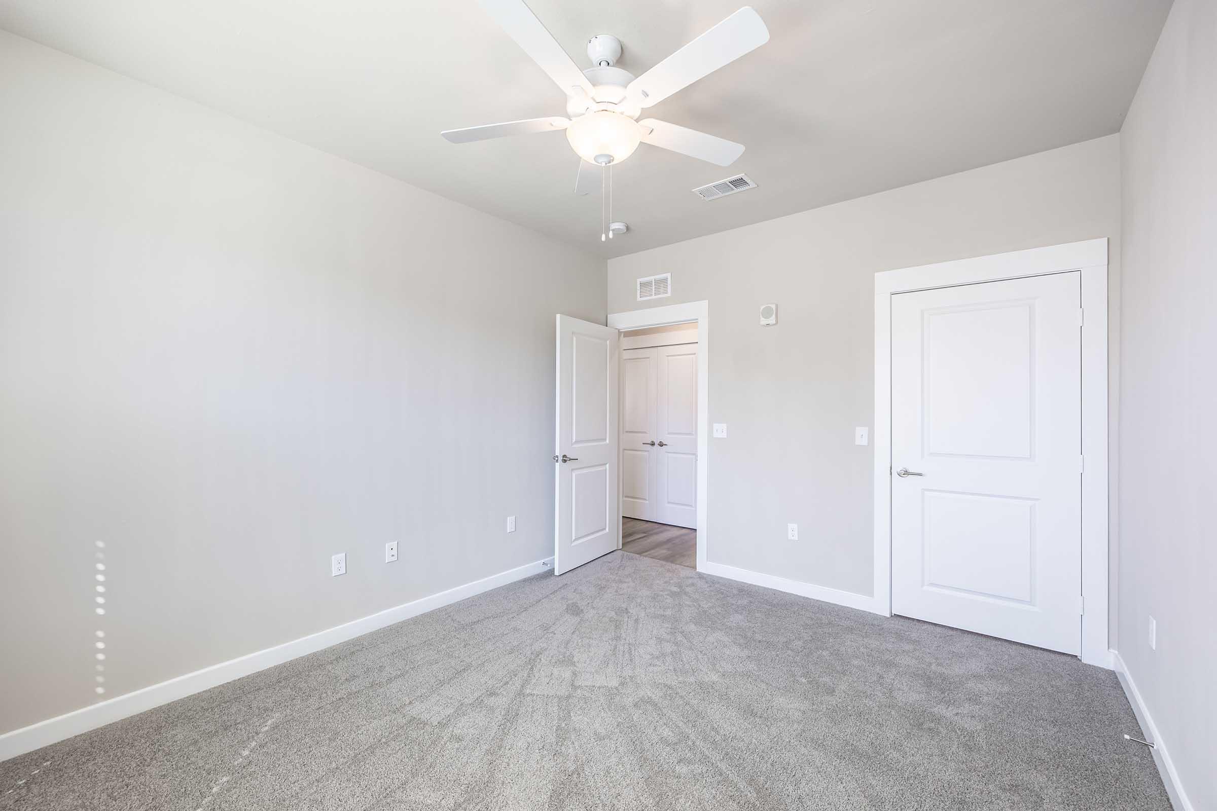 Empty room with light gray walls and a ceiling fan. The floor is covered in plush, light-colored carpet. There are two white doors, one partially open, leading to another area. Natural light streams in through a nearby window, creating a bright and airy atmosphere.