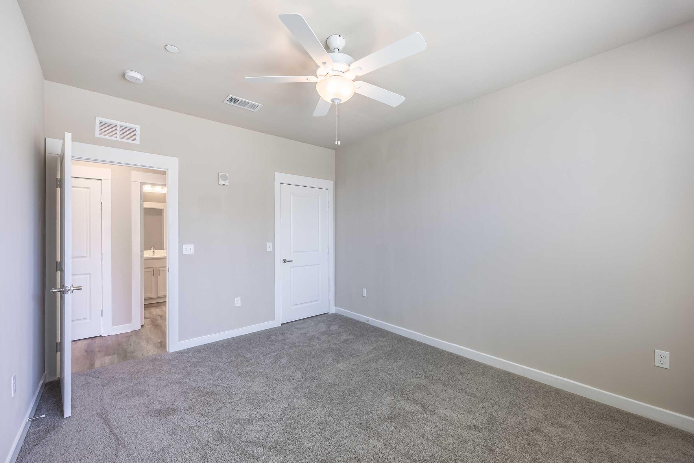 A well-lit, empty bedroom featuring gray carpet, soft walls, and a ceiling fan. A door leading to a bathroom is visible on the left, while the entrance door is slightly open on the right. The room has no furniture, creating a spacious and minimalist feel.