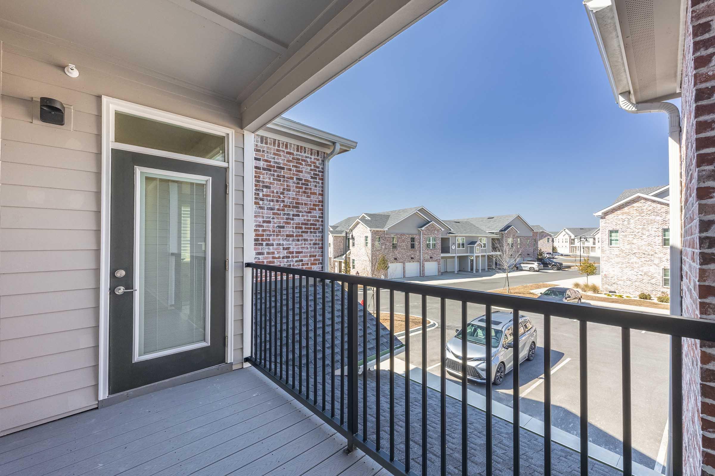 A view from a balcony showing a modern apartment complex, featuring brick and siding exteriors. The image captures a clear blue sky and a parking area with cars. The balcony is enclosed with metal railing, and there's a door leading into the apartment. The overall atmosphere is bright and inviting.