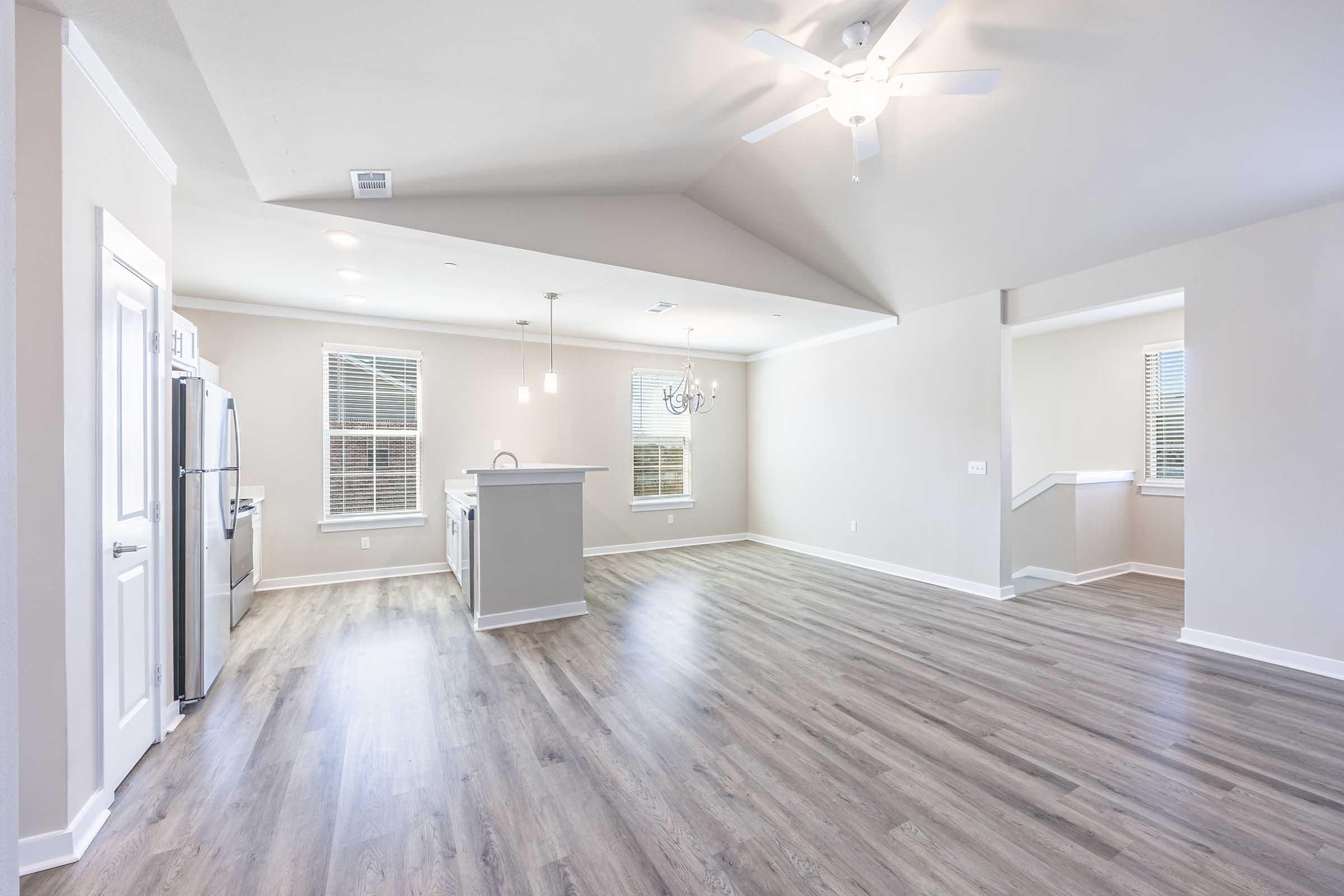 A spacious and modern living area featuring light wood flooring, white walls, and a ceiling fan. The room includes large windows for natural light, a contemporary chandelier, and an open kitchen with stainless steel appliances. Stairs lead to an upper level, enhancing the home's layout.