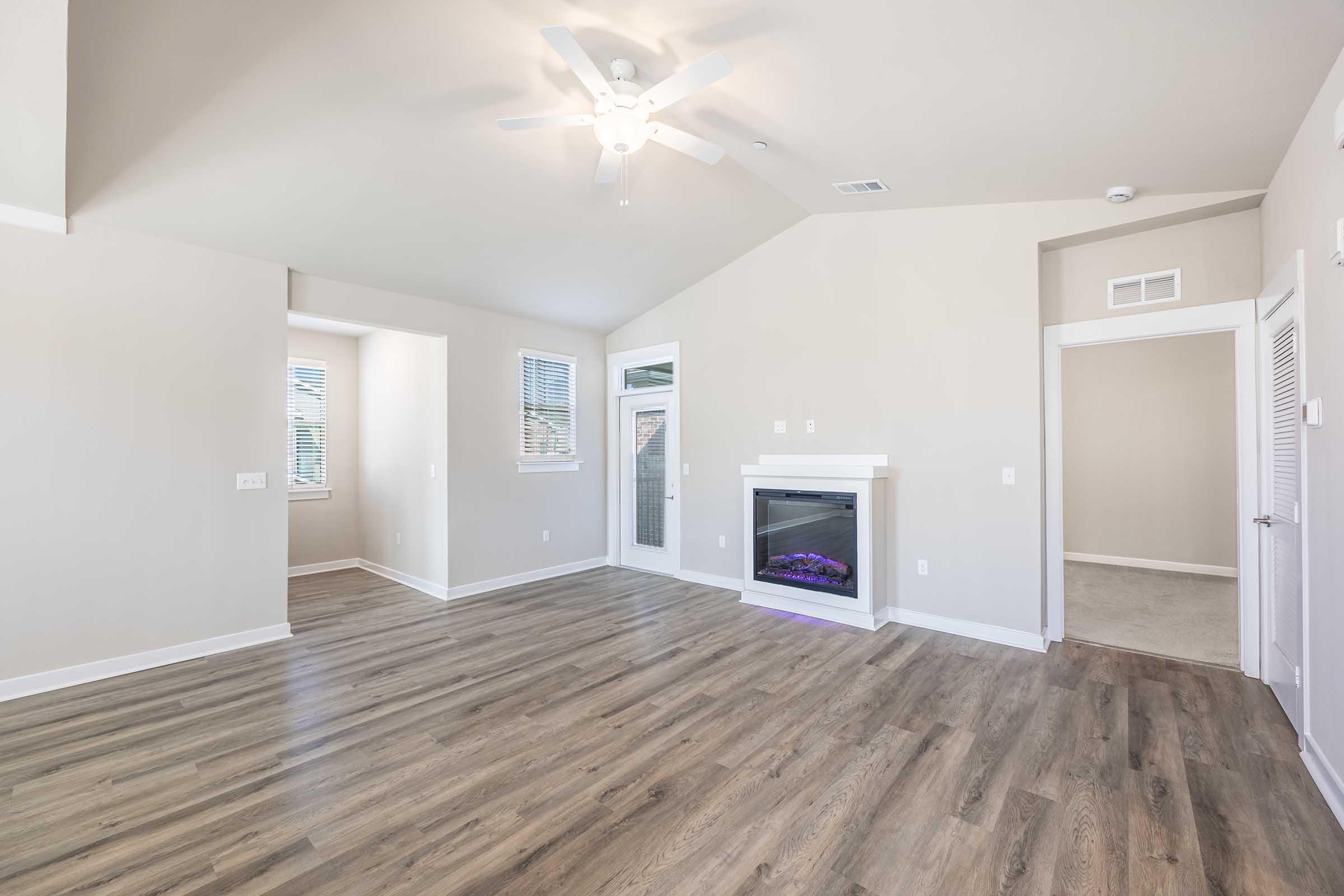 A spacious and well-lit living room with a ceiling fan, large windows, and light-colored walls. Features a white fireplace with an electric insert, modern laminate flooring, and doorways leading to adjoining rooms. The room is designed for comfort and openness.