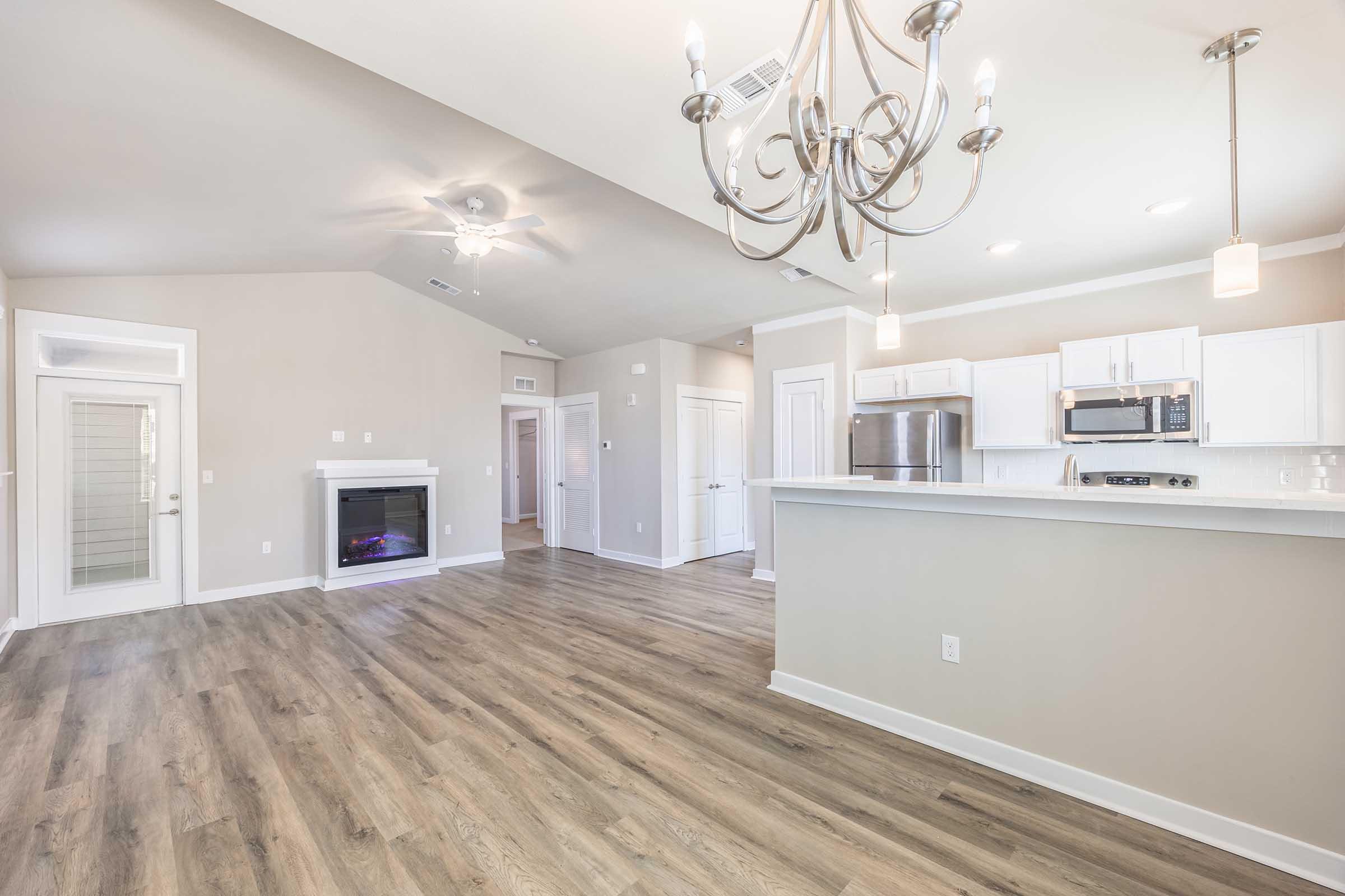 Spacious and modern living area featuring a stylish chandelier, large windows, and a cozy electric fireplace. The open floor plan connects to a contemporary kitchen with white cabinetry and stainless steel appliances. Light neutral colors enhance the inviting atmosphere, complemented by luxury vinyl flooring.