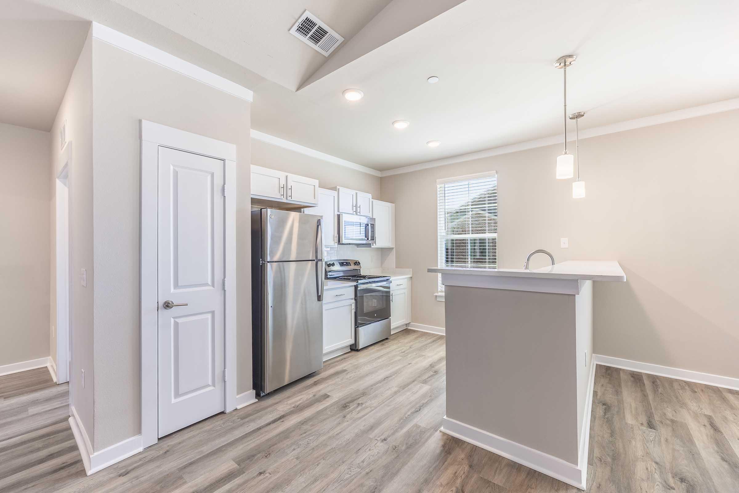 Modern kitchen featuring stainless steel appliances, including a refrigerator and stove. The space has white cabinetry, light-colored walls, and a wood-like floor. Natural light streams in through a window, and there are pendant lights above the countertop, creating a bright and inviting atmosphere.