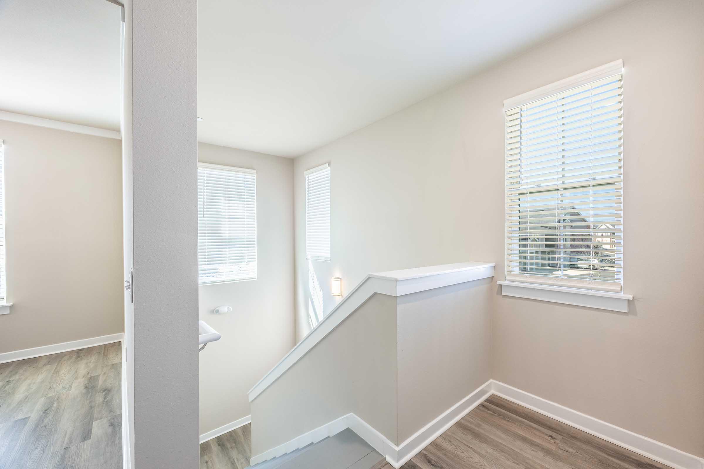 A well-lit interior space featuring a staircase leading up, with light-colored walls and hardwood flooring. There are two windows with blinds on the walls, allowing natural light to filter in. The overall ambiance is bright and modern, creating a welcoming atmosphere.