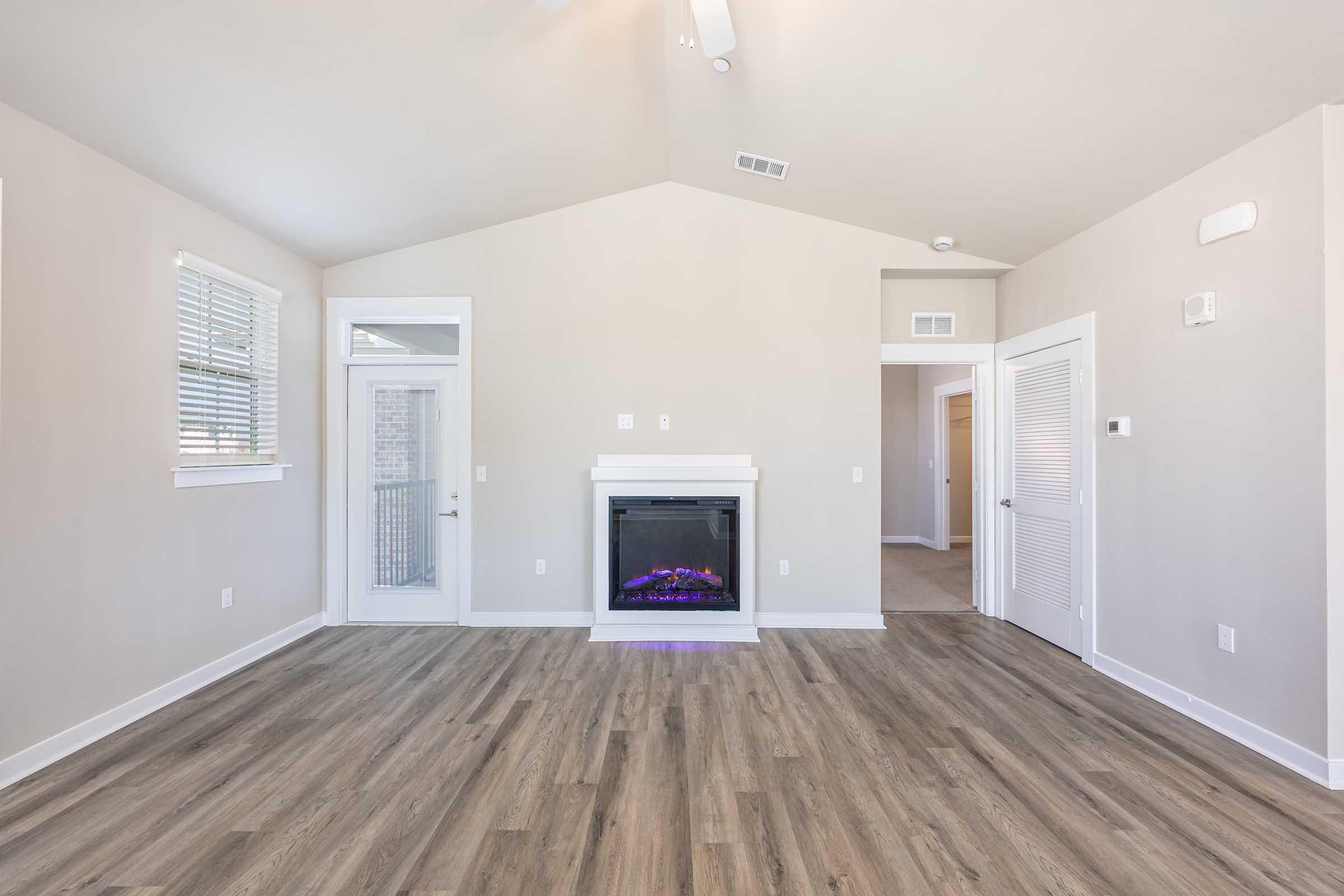Spacious room with light gray walls, large windows, and wood flooring. A modern electric fireplace is centered on one wall. There is a doorway leading to another room and a closet door visible. The ceiling is vaulted, adding to the airy feel of the space.
