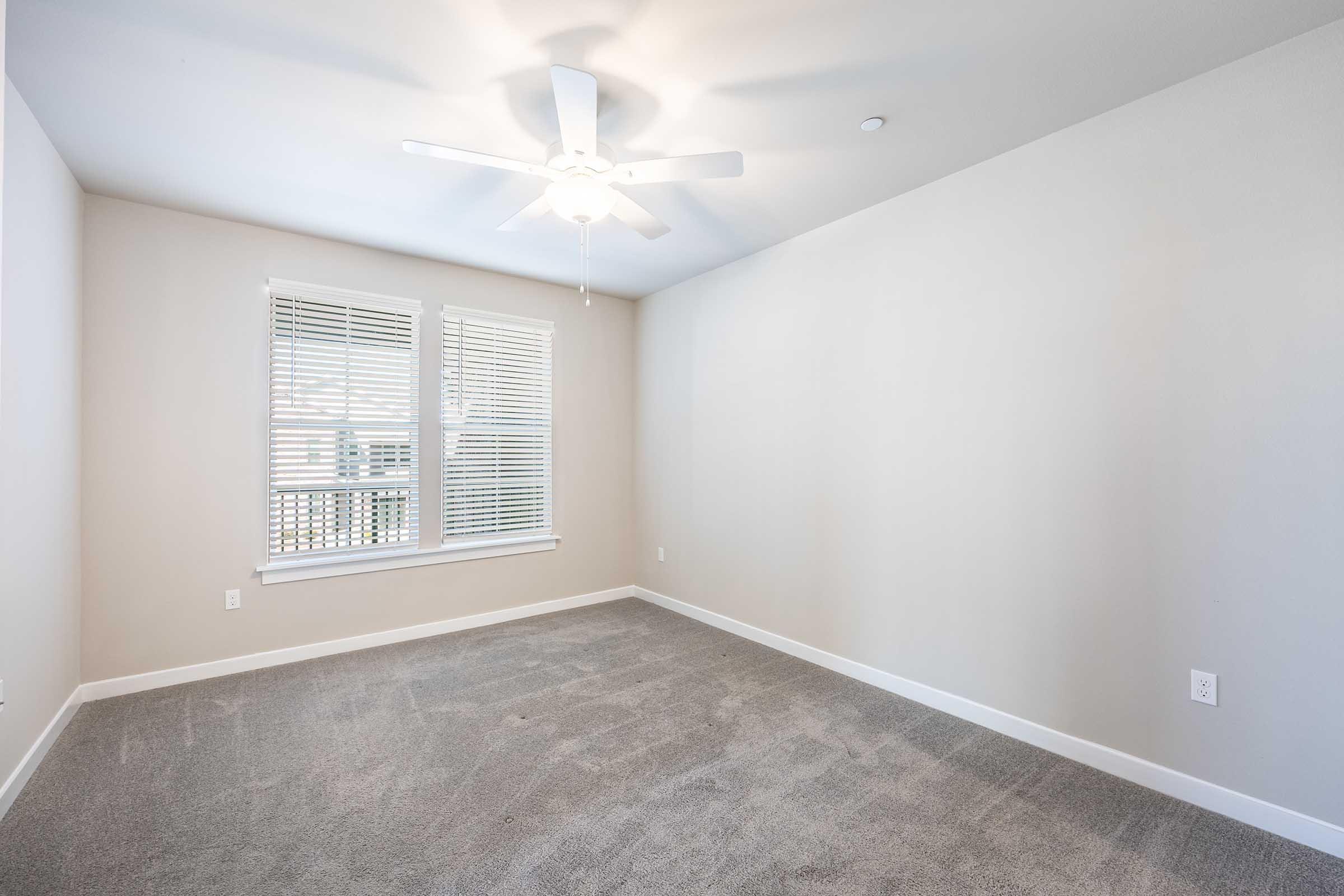 A well-lit, empty room with beige walls and light gray carpet. It features a ceiling fan and a window with white blinds that lets in natural light. The space is open and airy, ideal for decoration or furniture placement.