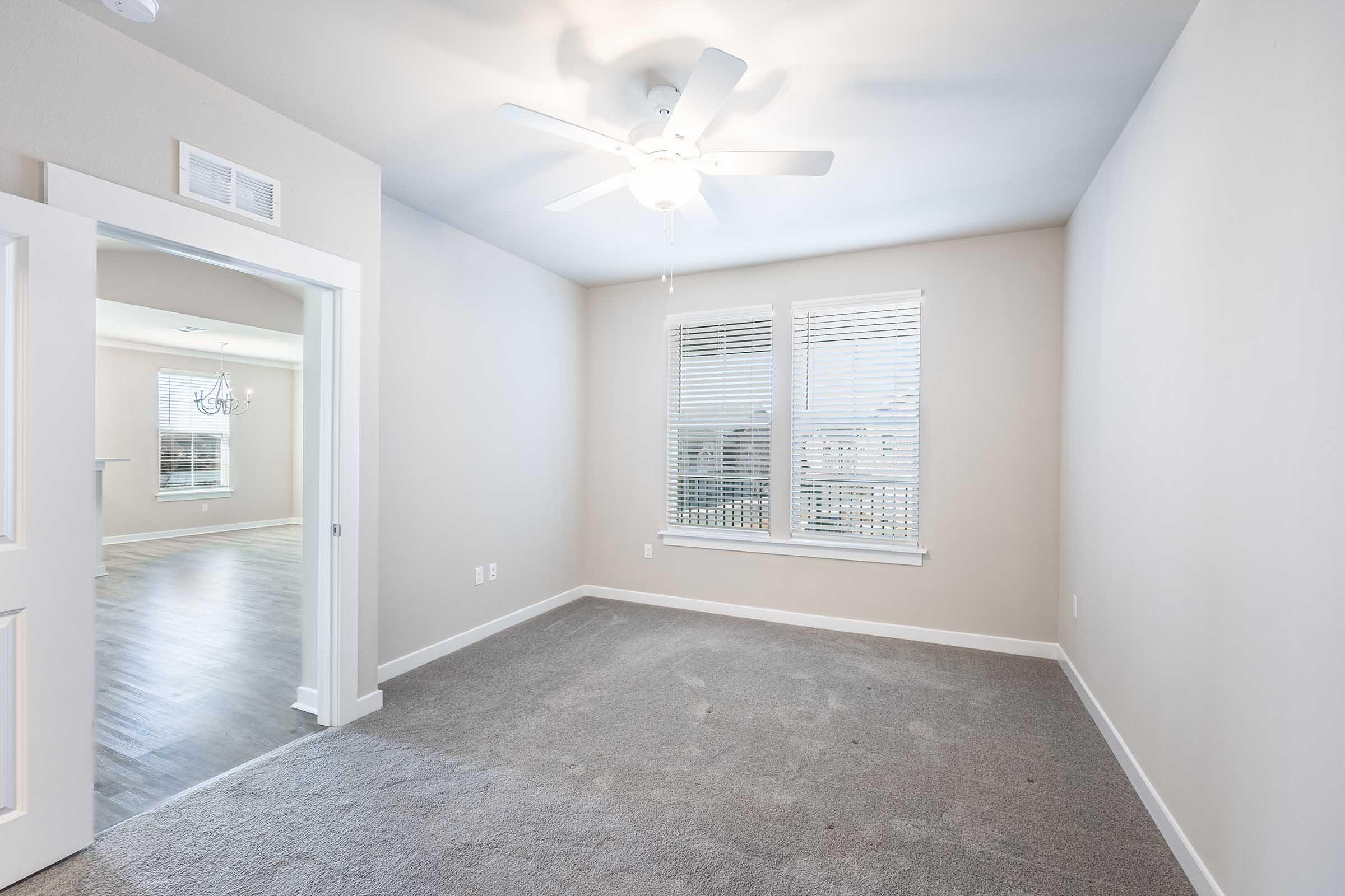 A minimalistic room with beige walls and gray carpeting. It features a ceiling fan and a window with blinds, allowing natural light to enter. A doorway leads to another room in the background. The space appears clean and ready for furnishing.