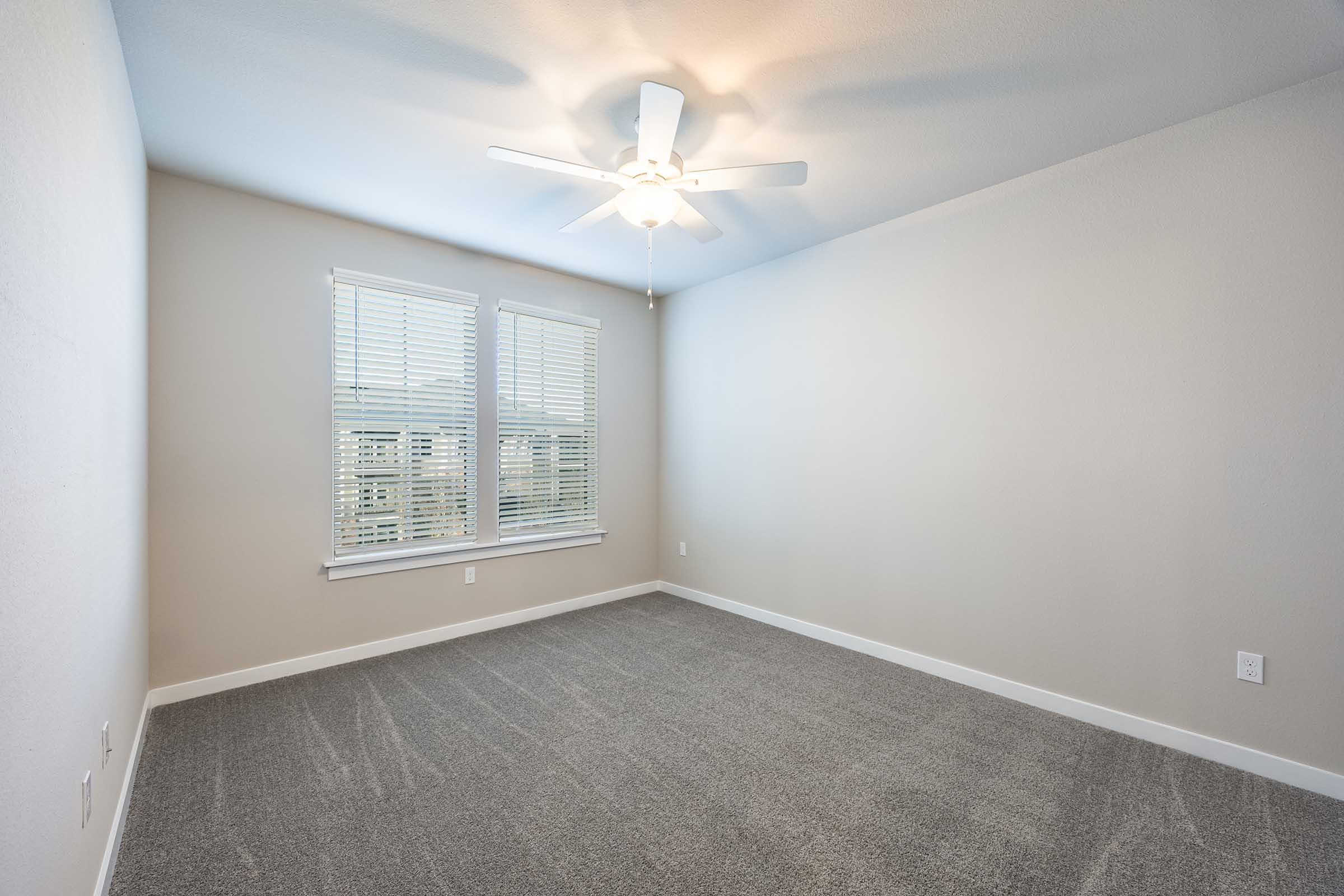 A bright, empty bedroom featuring light gray walls and carpet, a ceiling fan, and two large windows with white blinds offering a view outside. The room is well-lit, showcasing its spacious layout and clean design.