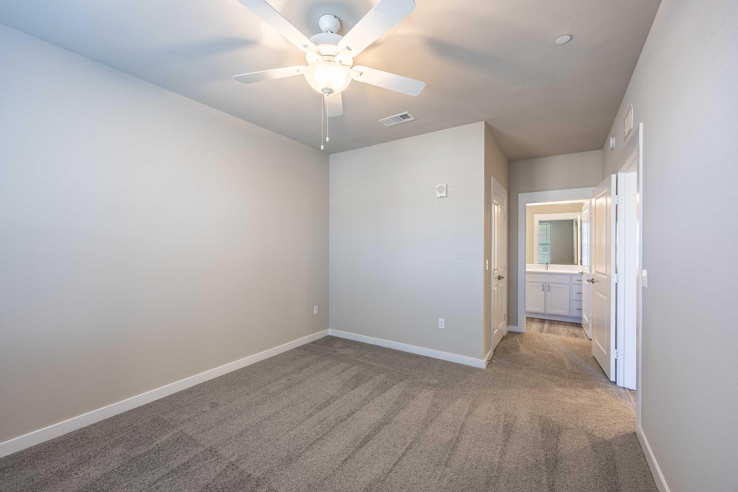A spacious, empty room with light gray walls and carpet. A ceiling fan is visible, and there is an open doorway leading to a bathroom area. Natural light comes in from a window outside the frame, enhancing the airy feel of the space.
