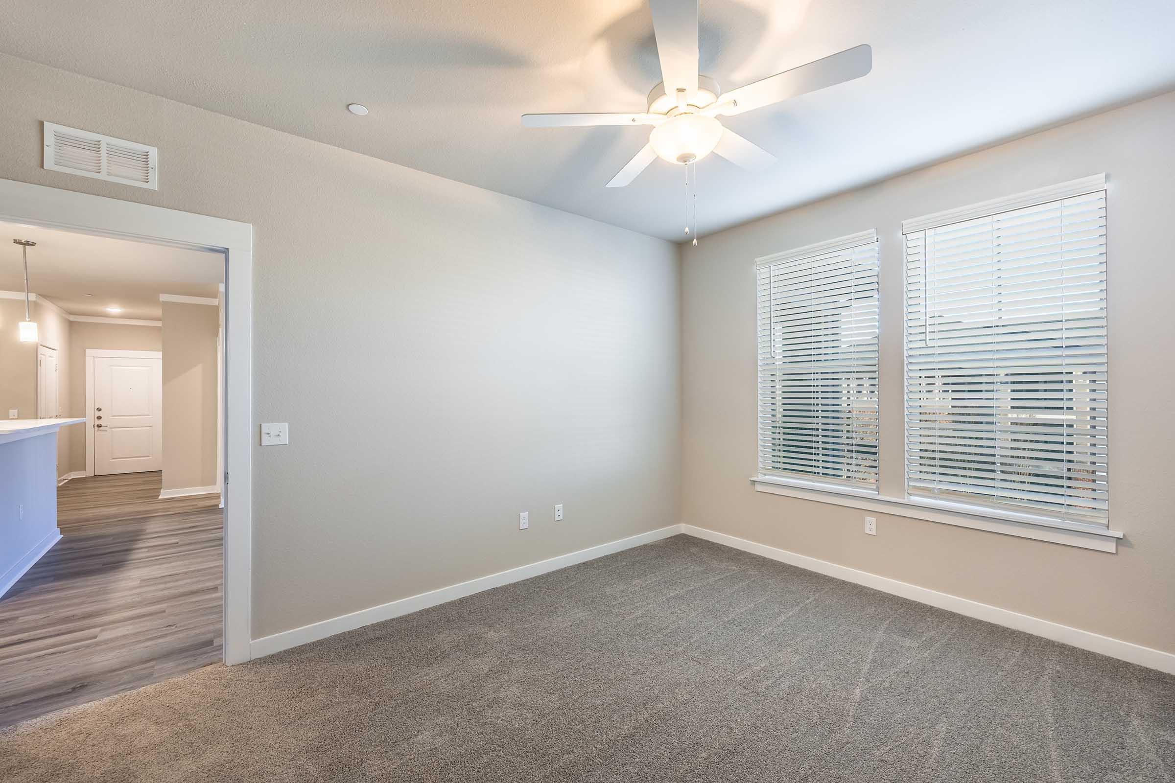 Empty room with light gray walls and carpet, featuring a ceiling fan and two large windows with blinds. A doorway on the left leads to a hallway, and a light-colored wall separates the room from an adjacent area. Natural light brightens the space.