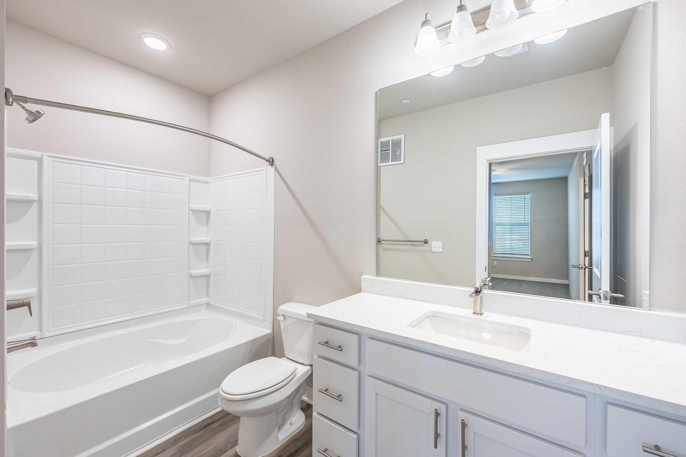 A modern bathroom featuring a white bathtub with a shower, a sleek vanity with a marble countertop and sink, a large mirror, and light fixtures. The walls are painted a soft beige, and there is a window in the background providing natural light.