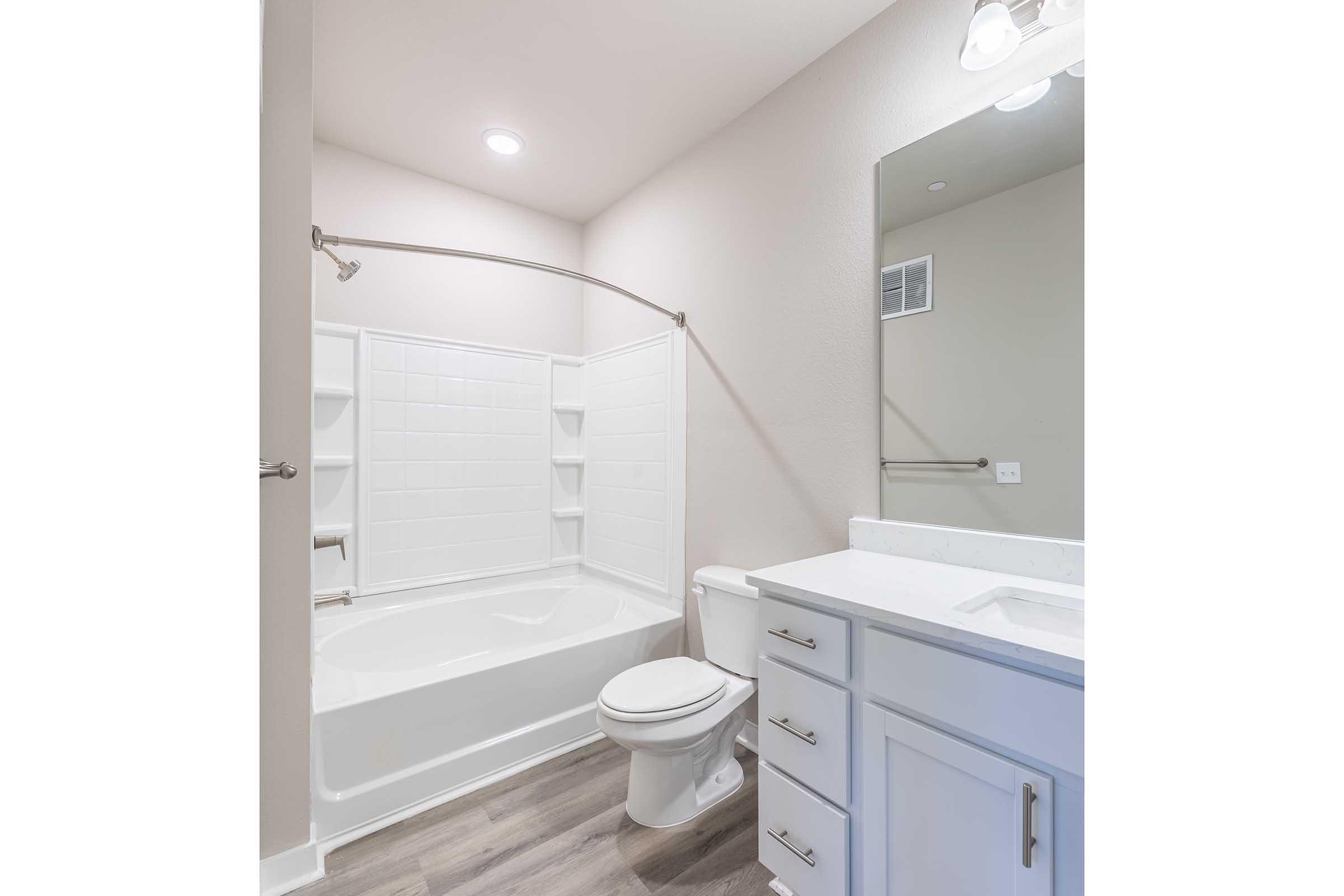 A modern bathroom featuring a bathtub with an attached shower, a white toilet, and a white vanity with drawers. Natural light illuminates the space, highlighting light-colored walls and wood-like flooring. A mirror is mounted above the sink, creating a clean and contemporary look.