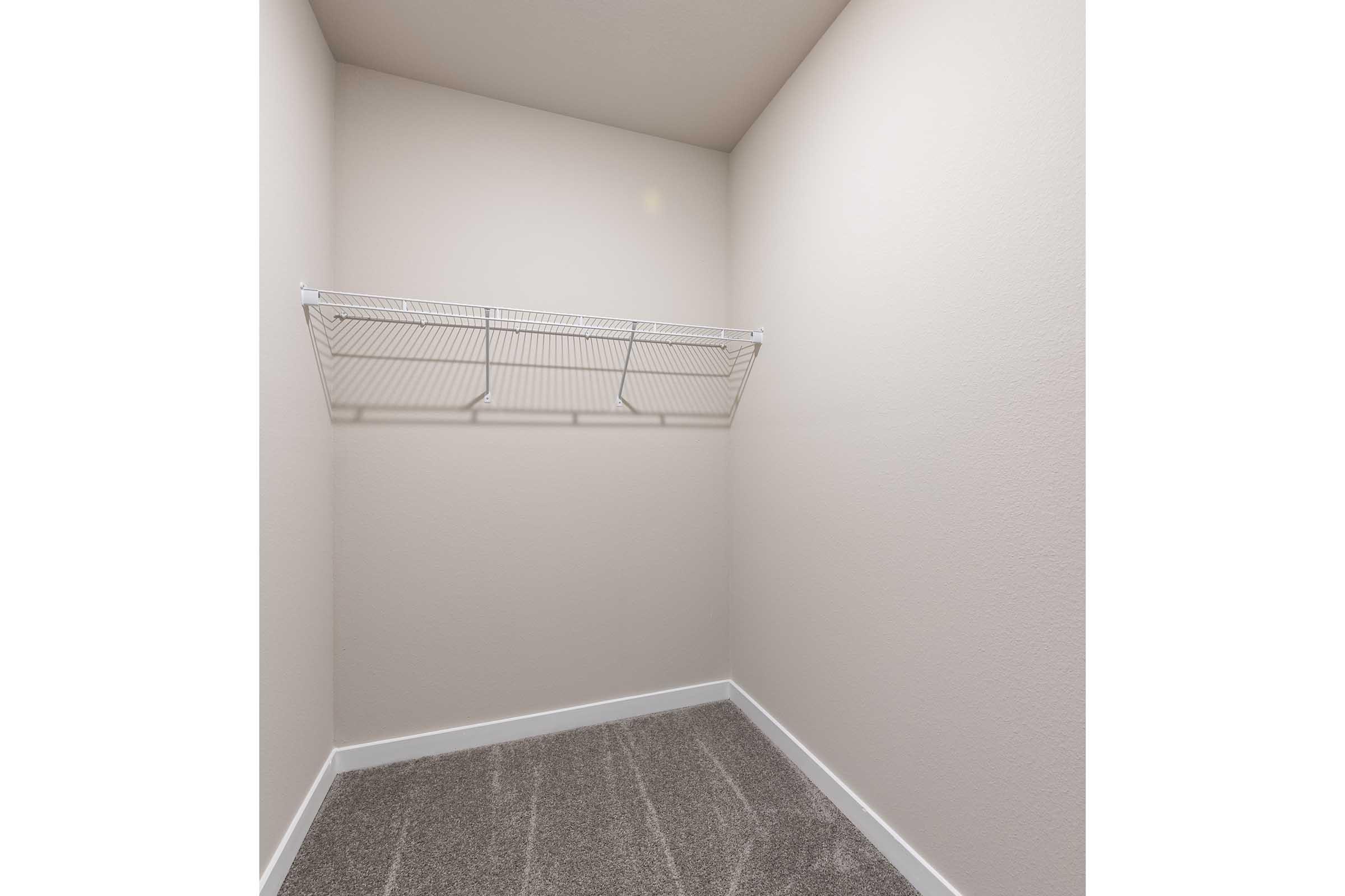 A small, empty closet with light-colored walls. The space features a wire shelf mounted on the back wall and a carpeted floor. The overall appearance is clean and minimalistic, suitable for storage.