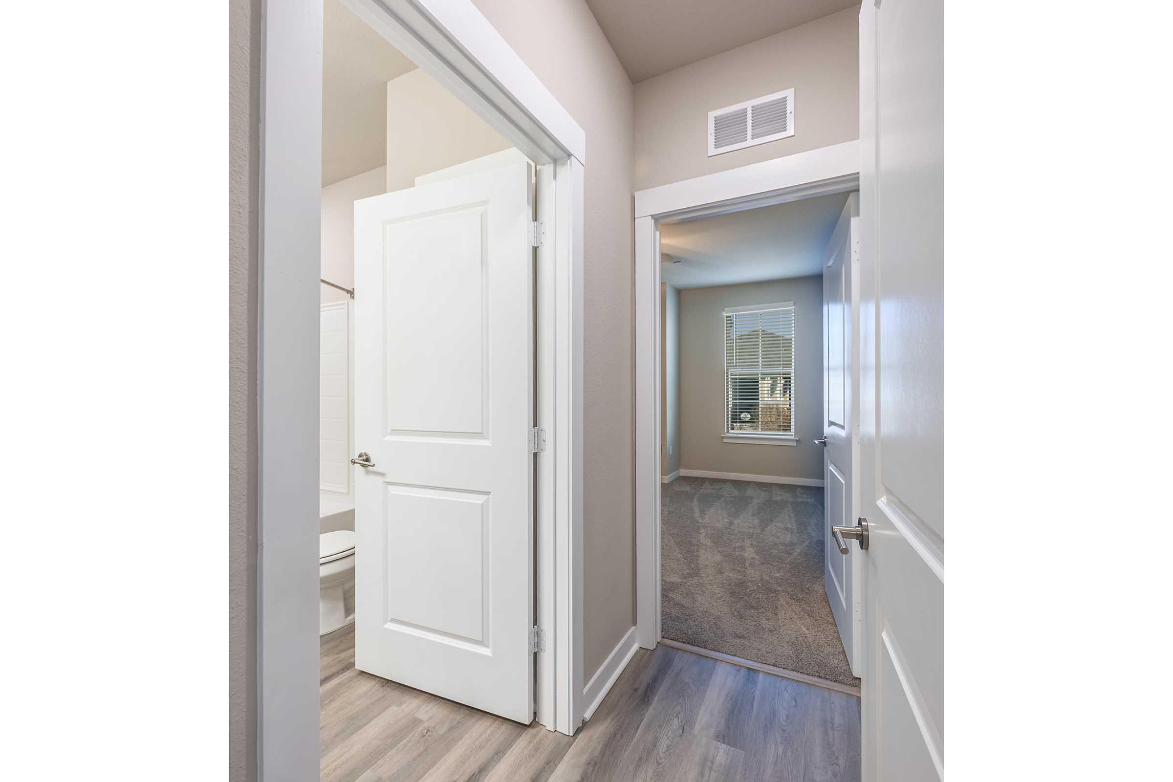 A hallway view showing an open bathroom door on the left with a white tiled wall and a bathtub visible. On the right, there is a door leading to another room with a carpeted floor and a window letting in natural light. The walls are painted in a neutral tone.
