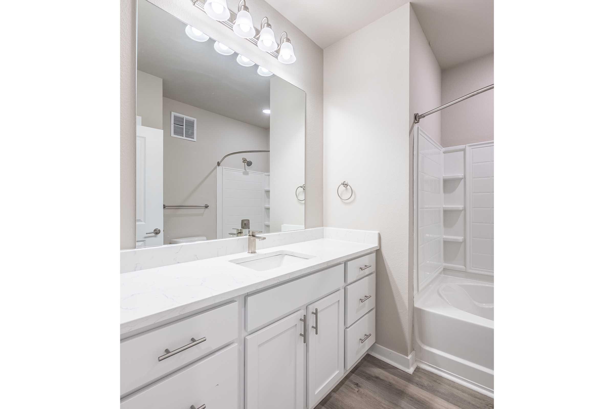 Modern bathroom featuring a white countertop with a sink, a large mirror above, and contemporary lighting fixtures. The bathroom includes a shower with a glass door and a bathtub. Neutral-colored walls and light wooden flooring complete the design, creating a clean and spacious atmosphere.