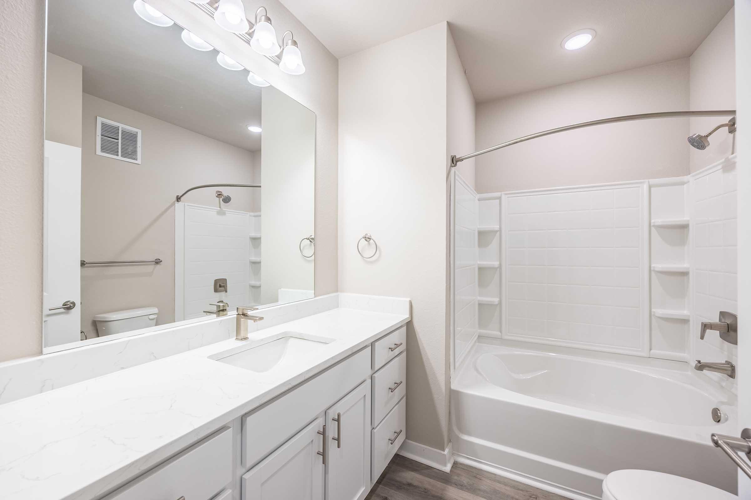 A modern bathroom featuring a white vanity with dual sinks, a large mirror, and bright lighting. The layout includes a shower-tub combo with a white surround and shelving, a toilet, and light-colored walls. The flooring is a wood-like material, creating a clean and contemporary look.