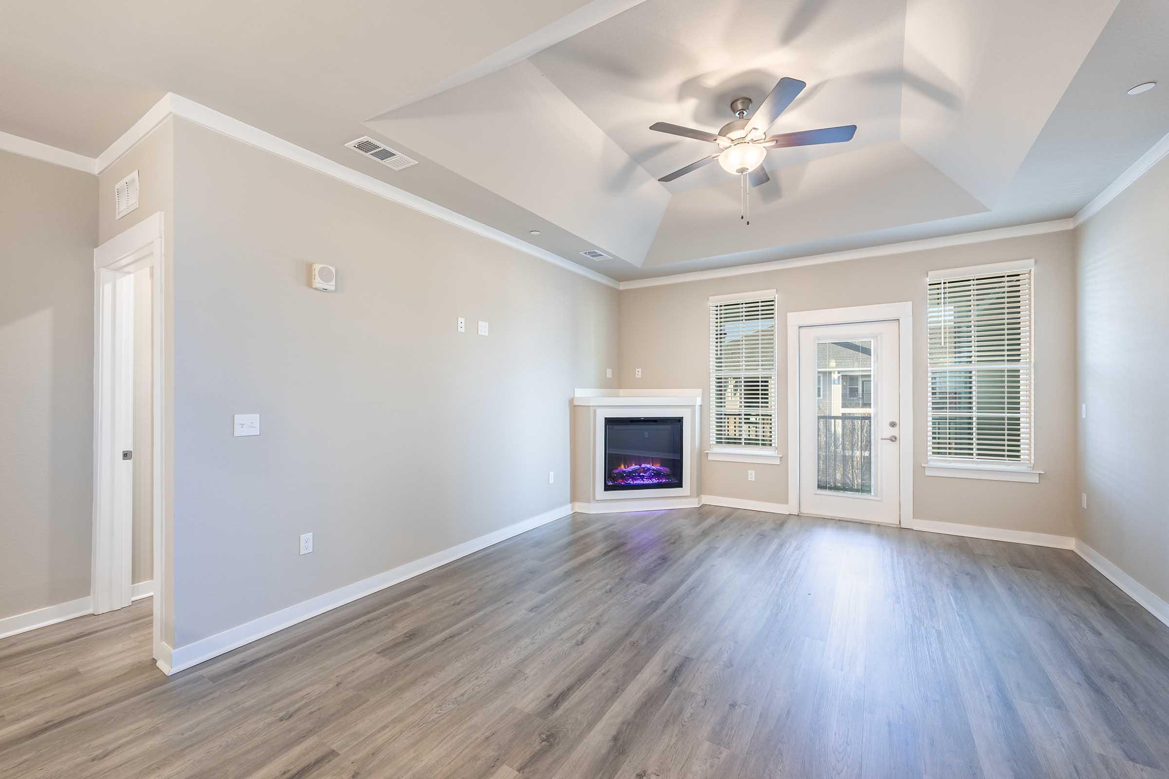 A spacious, modern living room featuring light-colored walls, wood-like laminate flooring, and a ceiling fan. There is an electric fireplace with a stone facade, large windows with blinds, and a door leading to an outdoor area, creating a bright and inviting atmosphere.