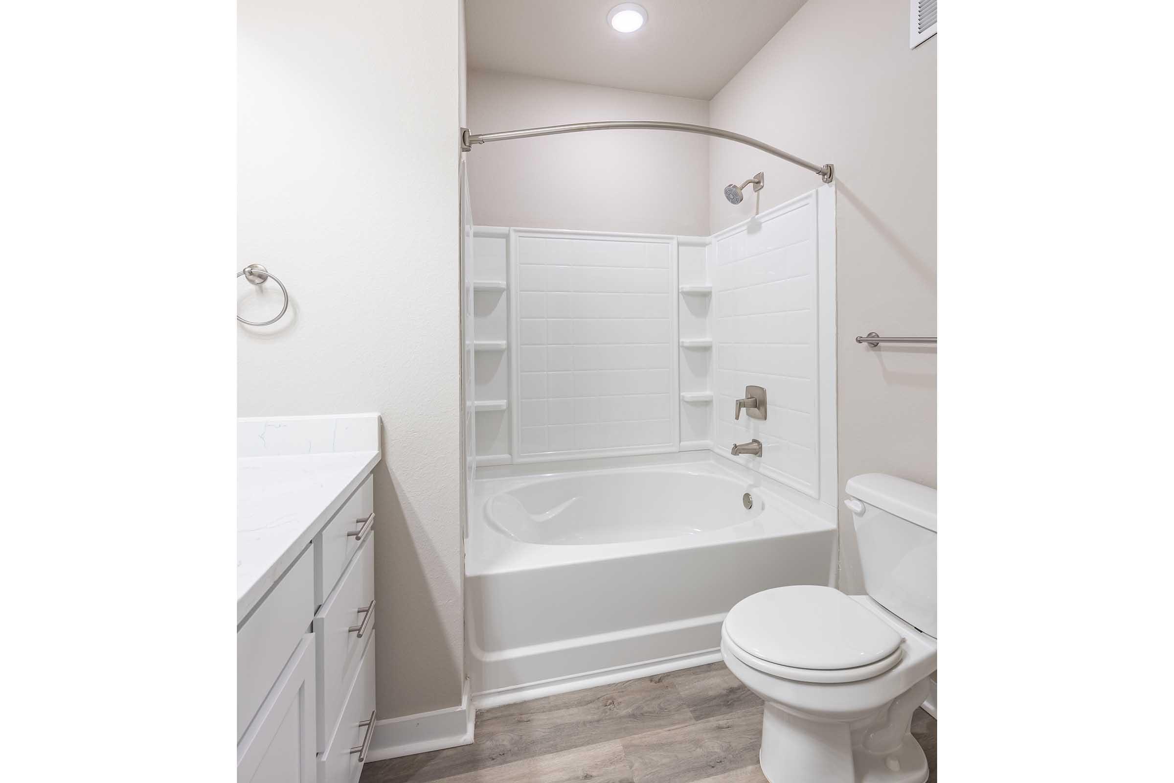 A clean and modern bathroom featuring a white bathtub with a curved shower curtain rod, built-in shelf for toiletries, a white toilet, and a vanity with drawers. The walls are light-colored, and the flooring has a wood-like finish, providing a bright and spacious feel.