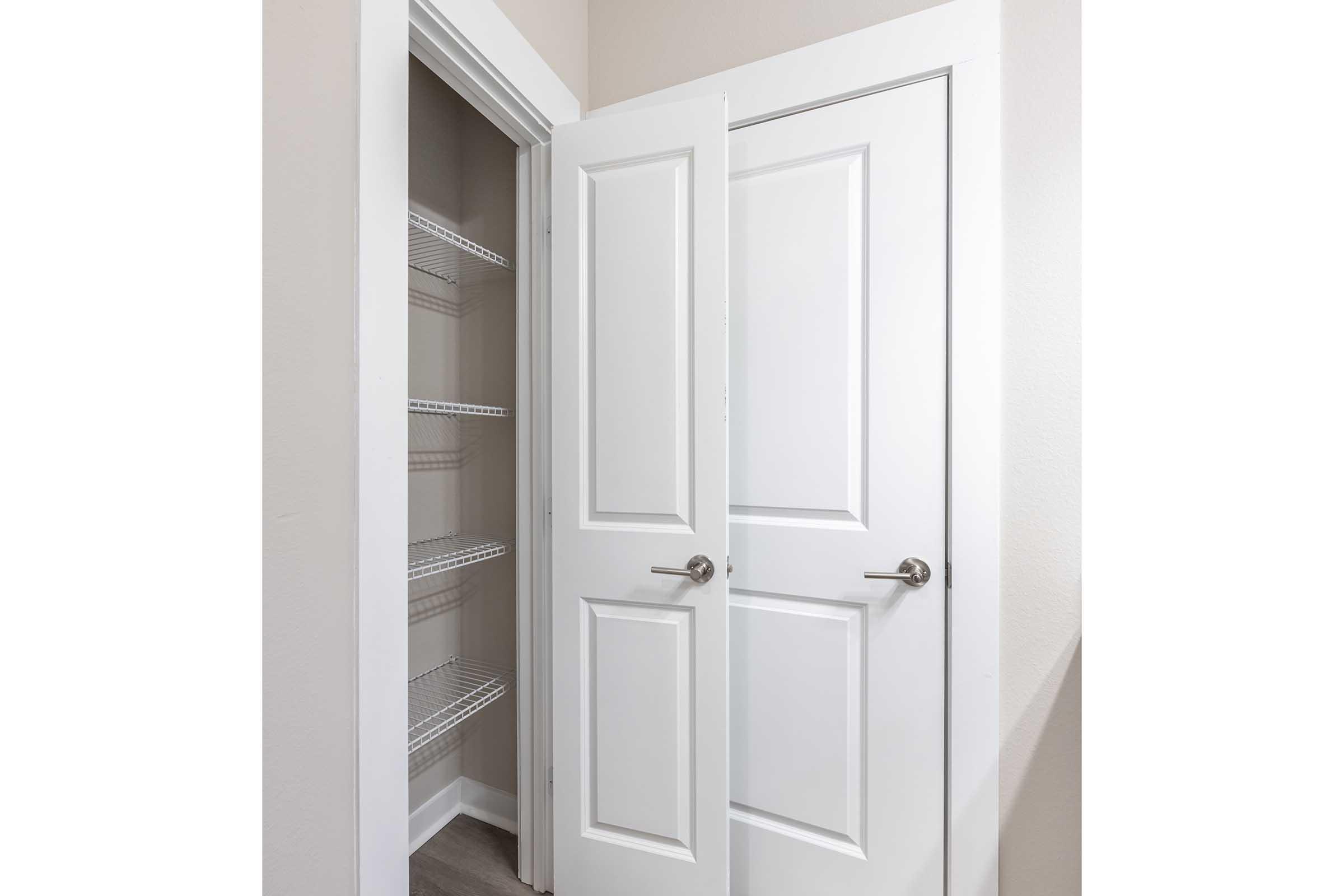 A partially open double door leads to a small closet with white wire shelves. The wall is a soft beige, and the floor is light gray. The closet appears organized and well-lit, showcasing a clean, modern aesthetic.