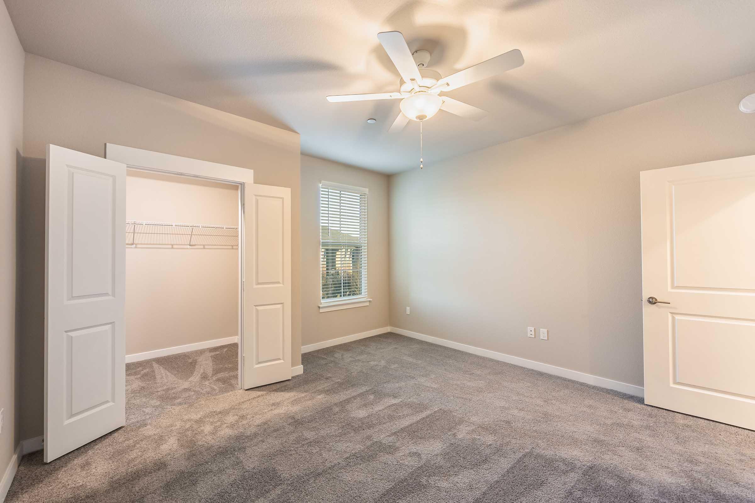 A well-lit, empty bedroom featuring light gray walls, a ceiling fan, and plush carpet. The room has two open white doors leading to a closet on one side and natural light streaming in from a window. The overall atmosphere is clean and spacious, suitable for personalization.