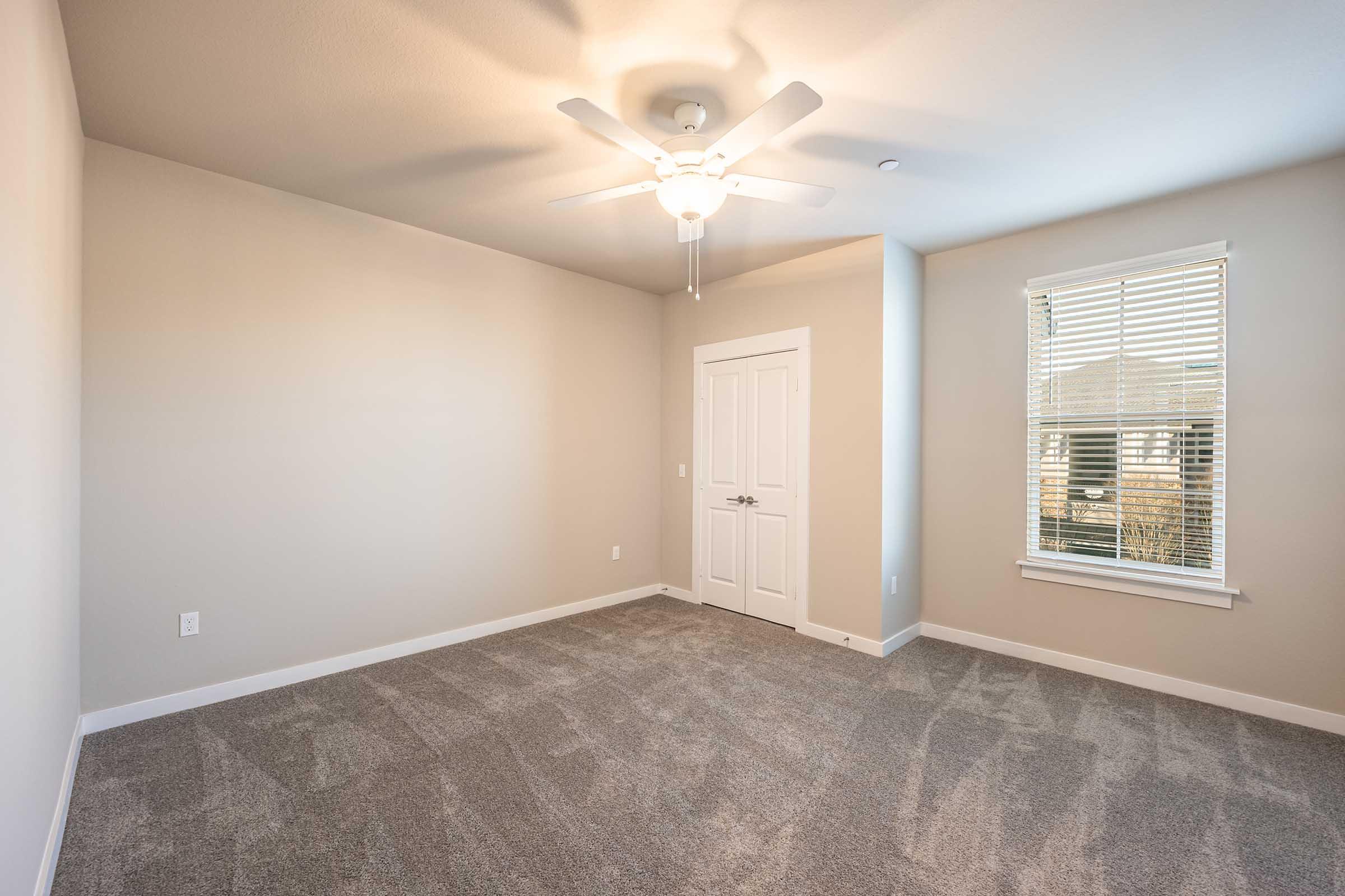 Empty room with light gray carpet and neutral walls. A ceiling fan is mounted in the center, and there is a window with blinds on the right side. A white door is visible on the left side of the image. The overall atmosphere is bright and inviting, with natural light coming through the window.