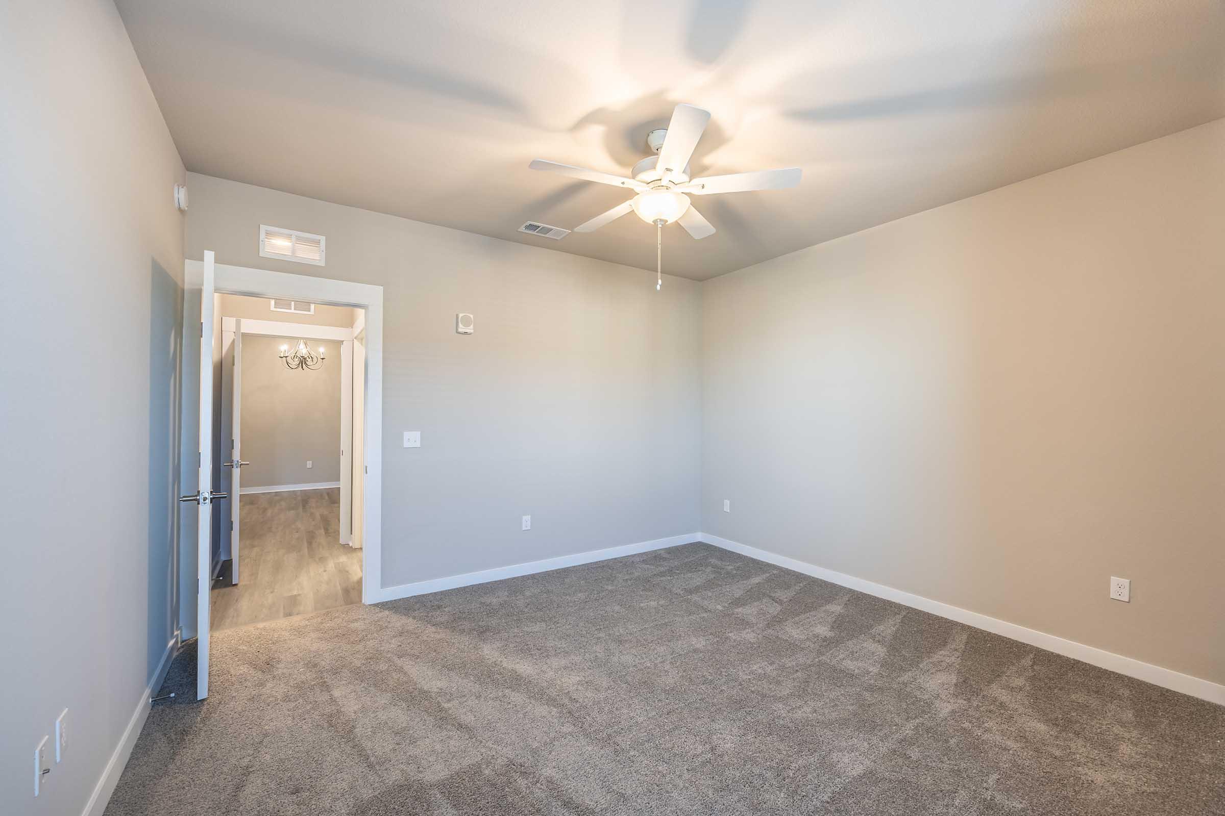 Empty room with light gray walls, a ceiling fan, and carpeted flooring. A doorway leads to another room in the background. The space is well-lit and features a simple, modern design without furniture or decorations.