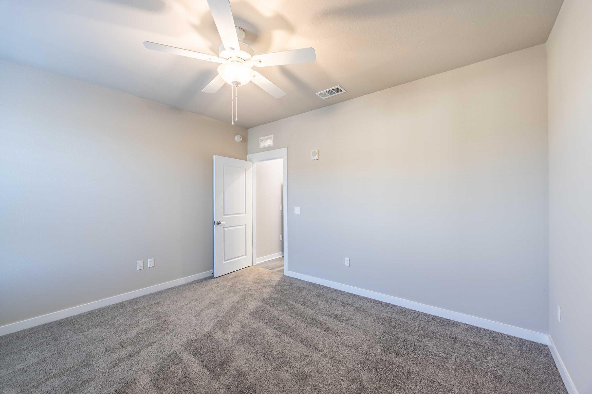 A spacious, empty room with light gray walls and a plush carpet. A ceiling fan is installed, and there is a white door leading to another area. Natural light enters from a nearby entrance. The overall atmosphere is bright and inviting, suitable for various uses.