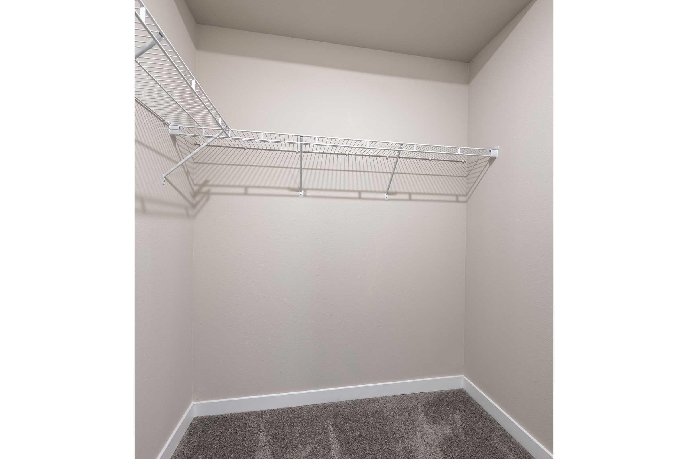 A vacant closet with light-colored walls and a gray carpeted floor. There is a wire shelf mounted on the wall, providing storage space, but no items or personal belongings are present. The overall appearance is clean and minimalistic.