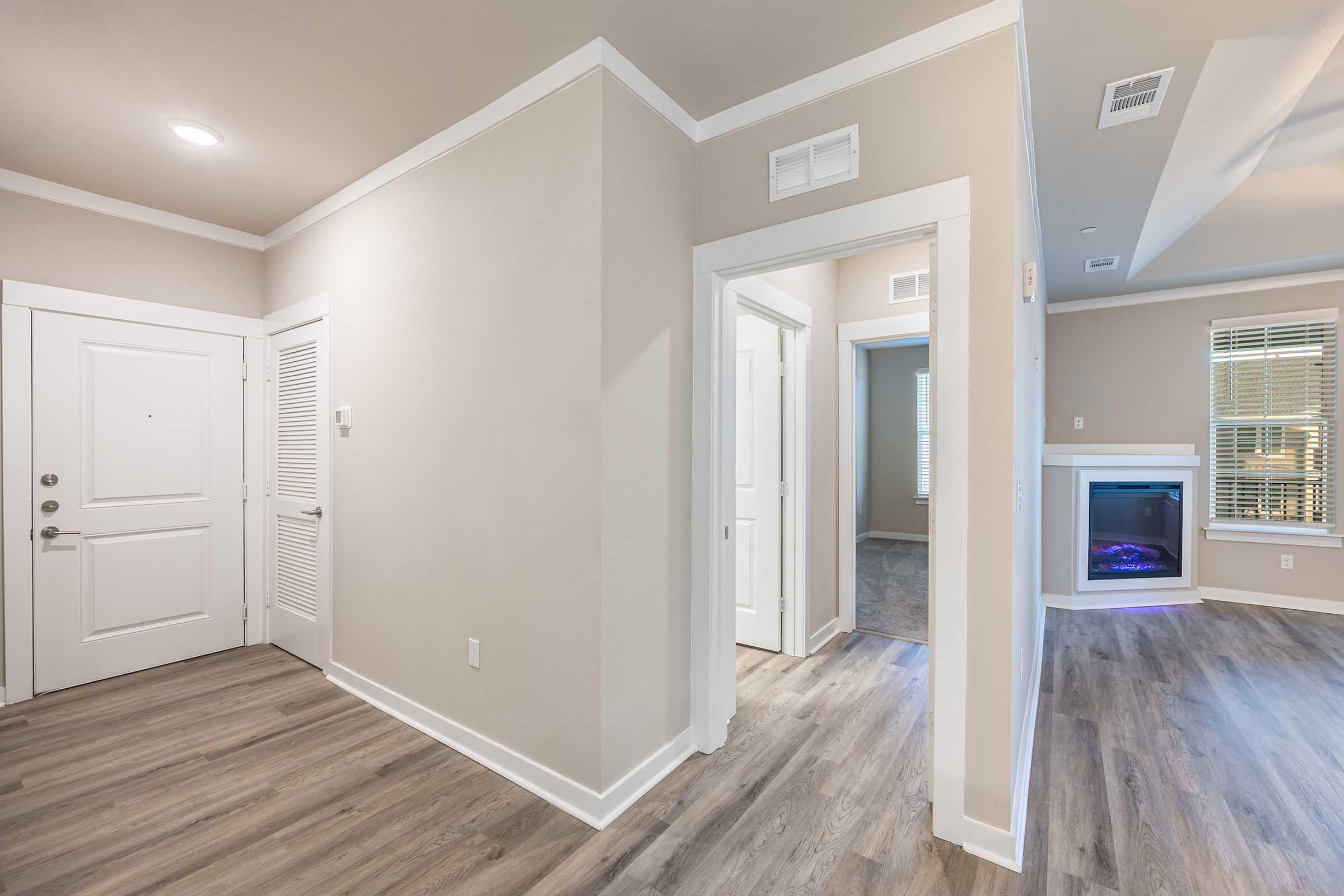 Interior view of a modern apartment entryway featuring light-colored walls, a partially visible door to the left, and an adjacent room with a window. The flooring is wooden, and there is a corner electric fireplace with blue lighting. The space is well-lit and has a welcoming ambiance.