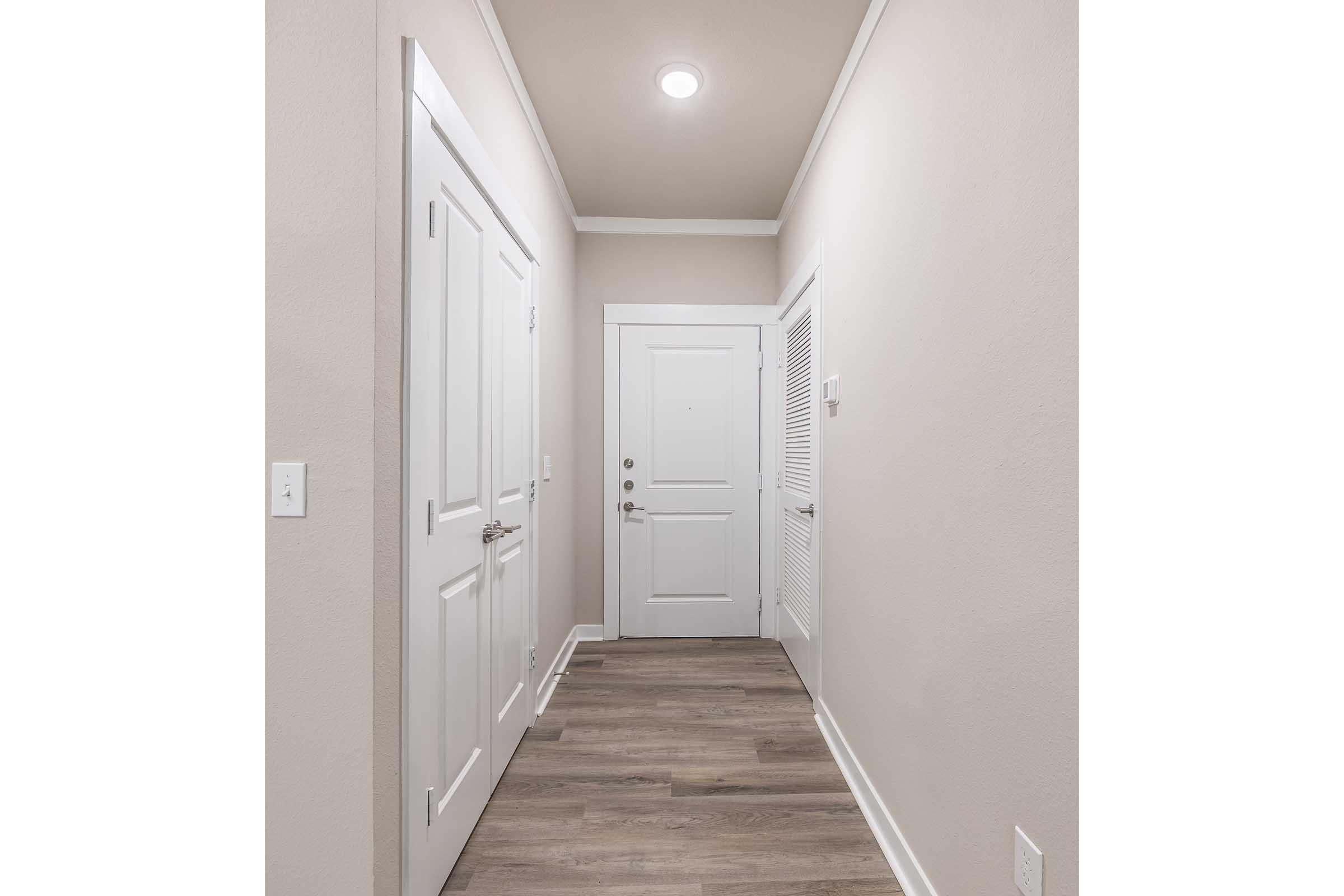 A well-lit hallway featuring light gray walls and a wood-like floor. There are two doors on the left, one with a handle and the other with a vent. A third door is at the end of the hallway, leading outside. The ceiling is white with a central light fixture.