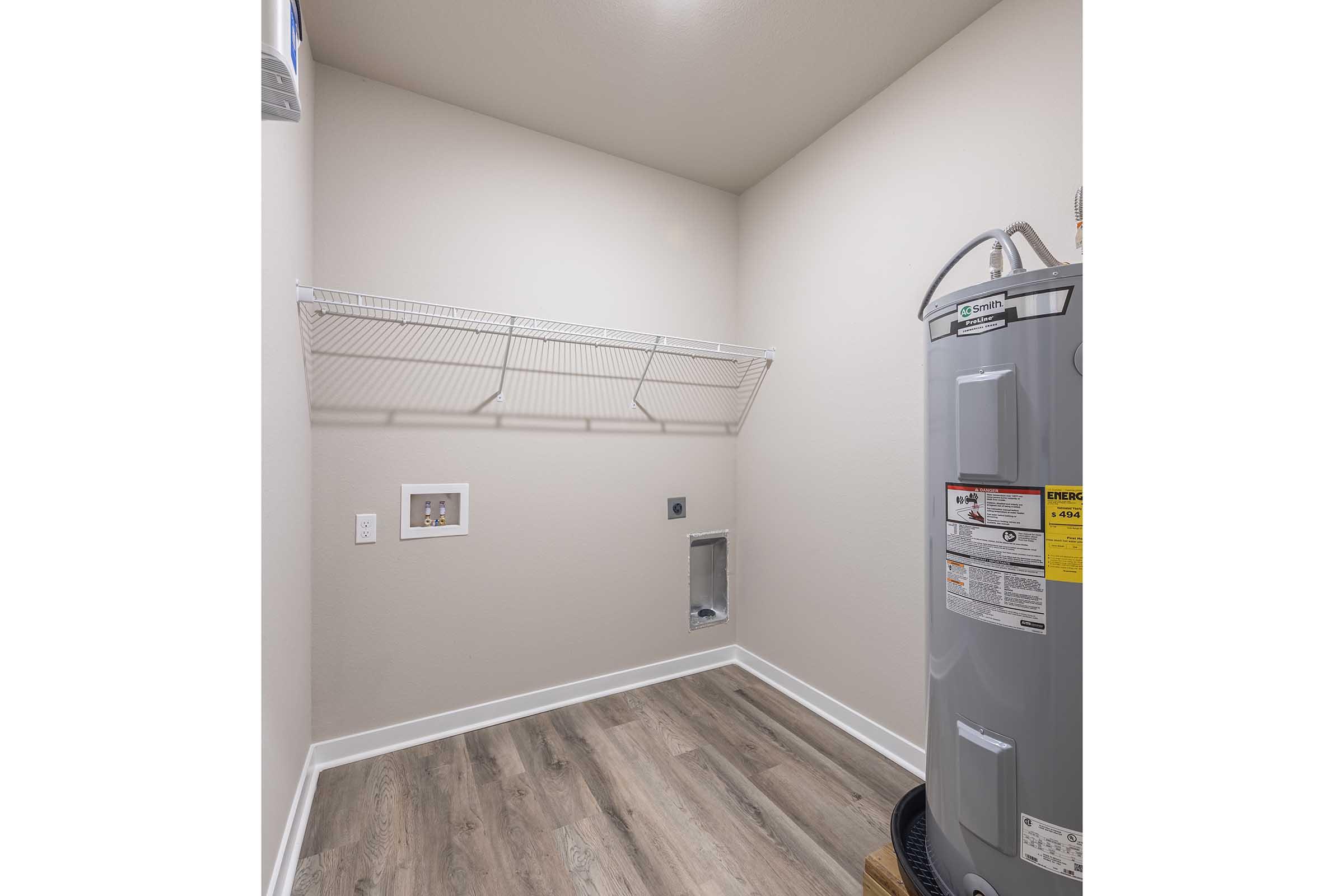 A spacious laundry room featuring a gray water heater, a wall-mounted wire shelf for storage, and a vent. The floor is covered with light wood-like laminate, and the walls are painted a neutral color, creating a clean and organized space.
