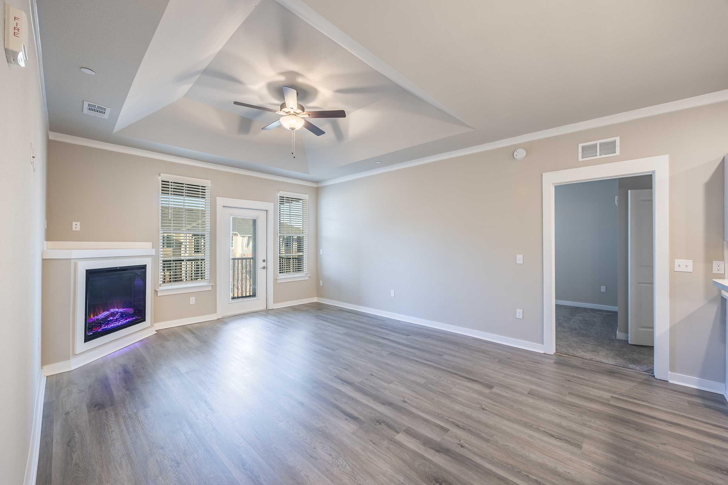 A spacious living room showcasing light-colored walls and hardwood flooring. Features include a ceiling fan, modern fireplace with purple lighting, large windows allowing natural light, and a door leading to a balcony. An adjacent doorway leads to another room, enhancing the open floor plan.