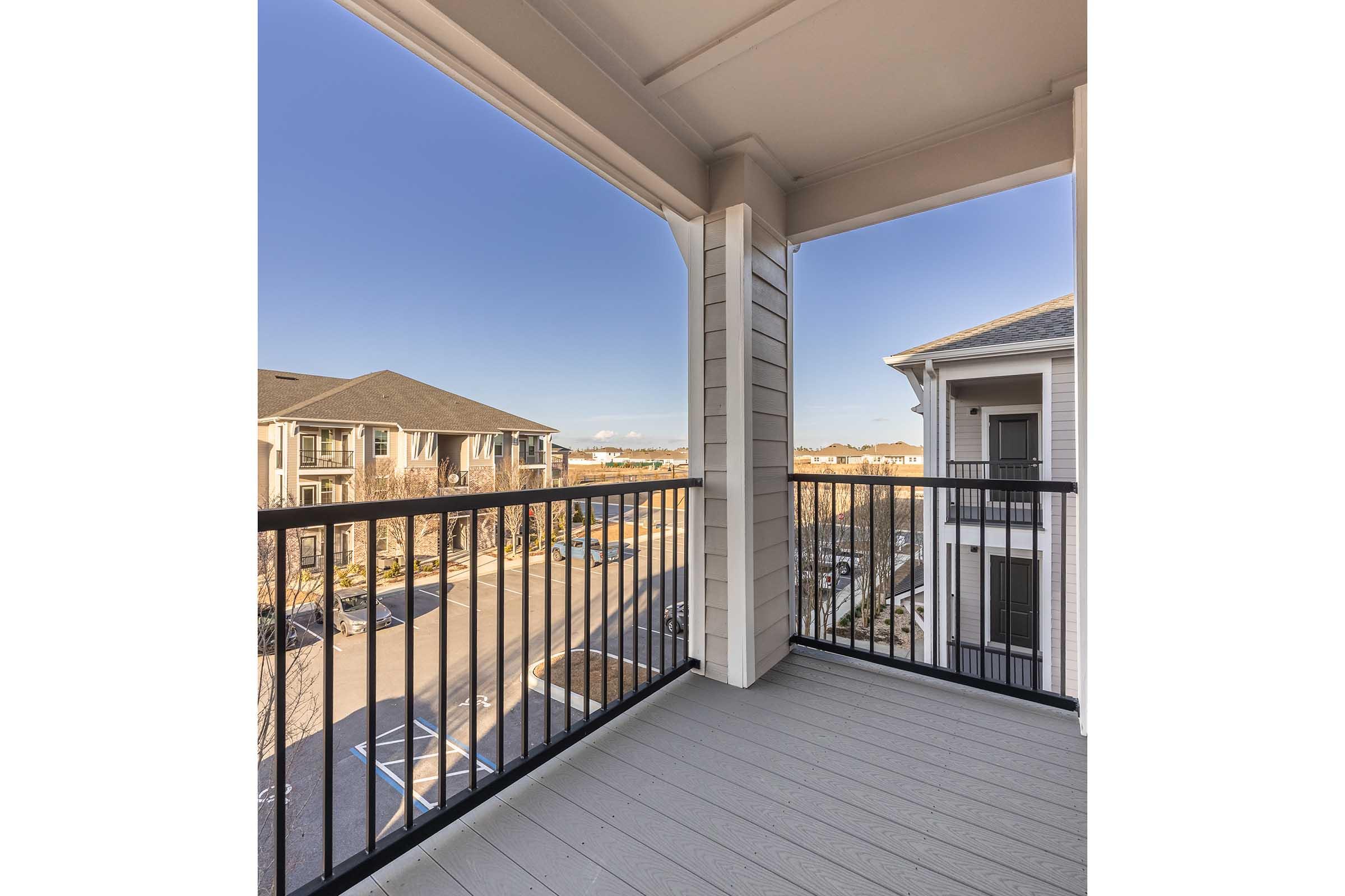 View from a balcony showcasing a clear blue sky and a neighborhood below. The balcony features a black railing and light-colored flooring. In the distance, several multi-story apartment buildings and a parking lot are visible, indicating a residential area.
