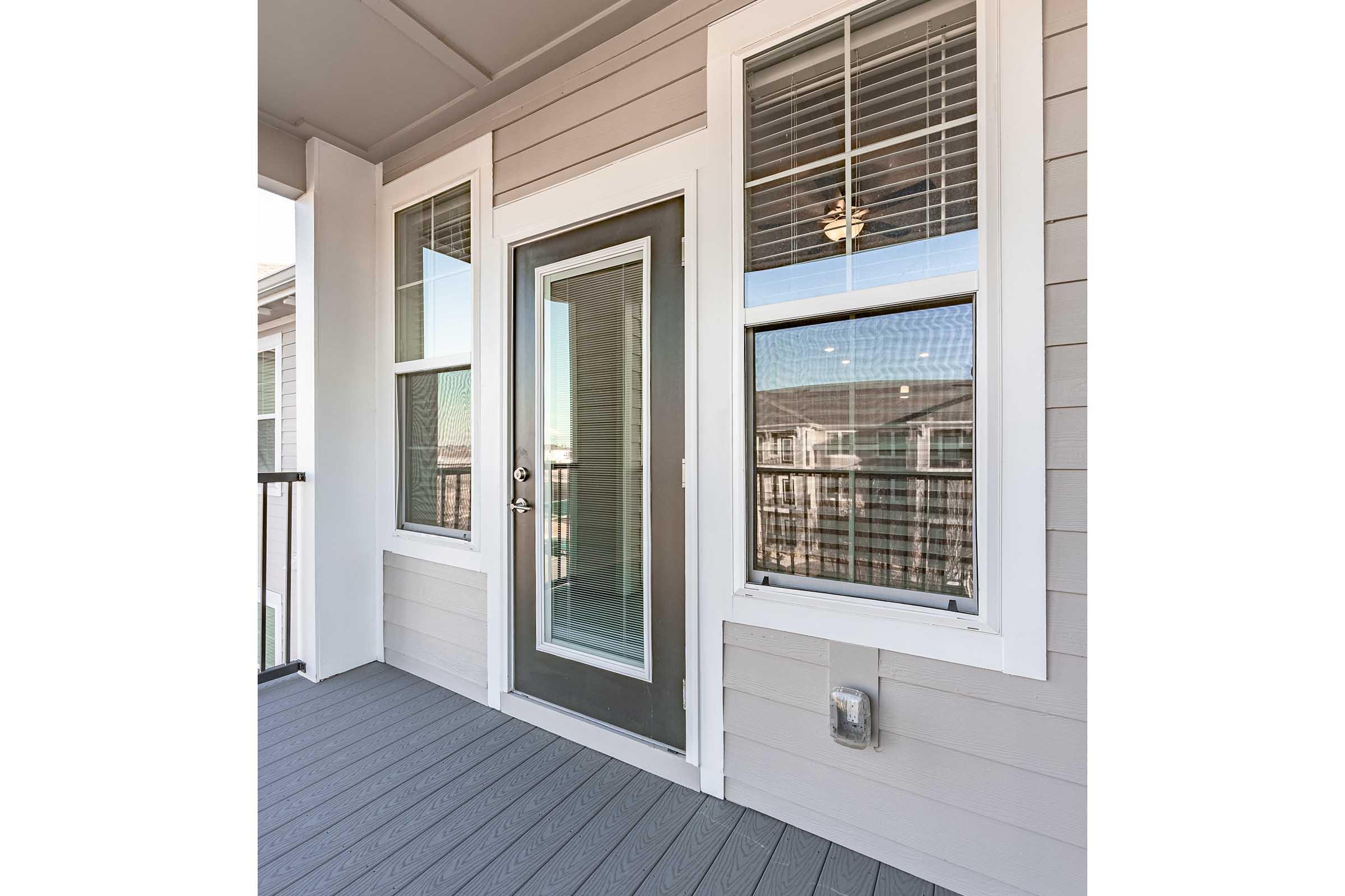 A modern entrance door framed by large windows on a covered porch. The door features a glass panel and is flanked by two windows that allow natural light. The porch floor is gray, matching the wall panels, creating a clean and contemporary aesthetic.