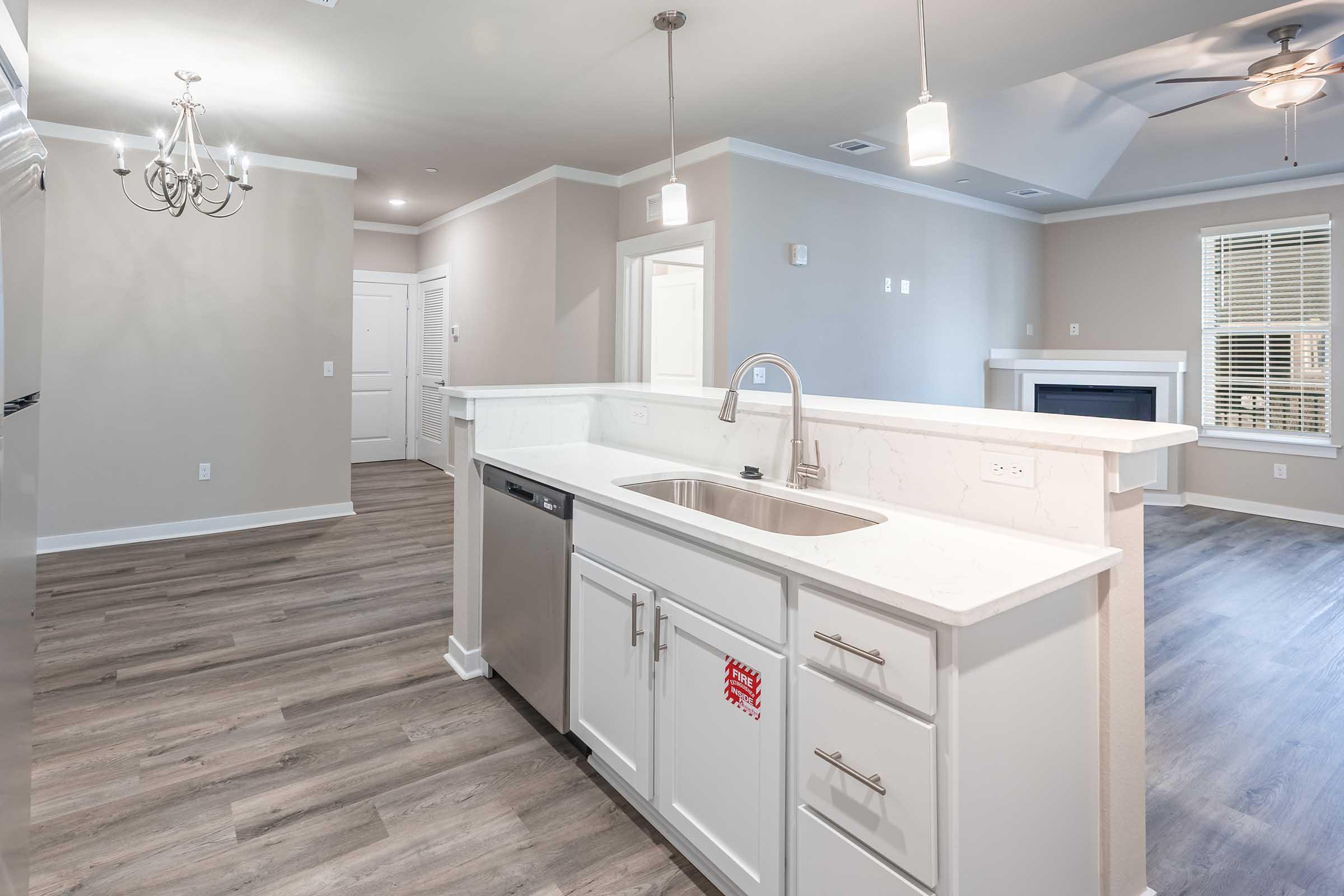 Modern kitchen and living area in a stylish apartment. The kitchen features a white countertop, stainless steel sink, and dishwasher. The open layout leads to a spacious living room with a ceiling fan and large windows, allowing natural light to fill the space. Light hardwood flooring enhances the contemporary design.