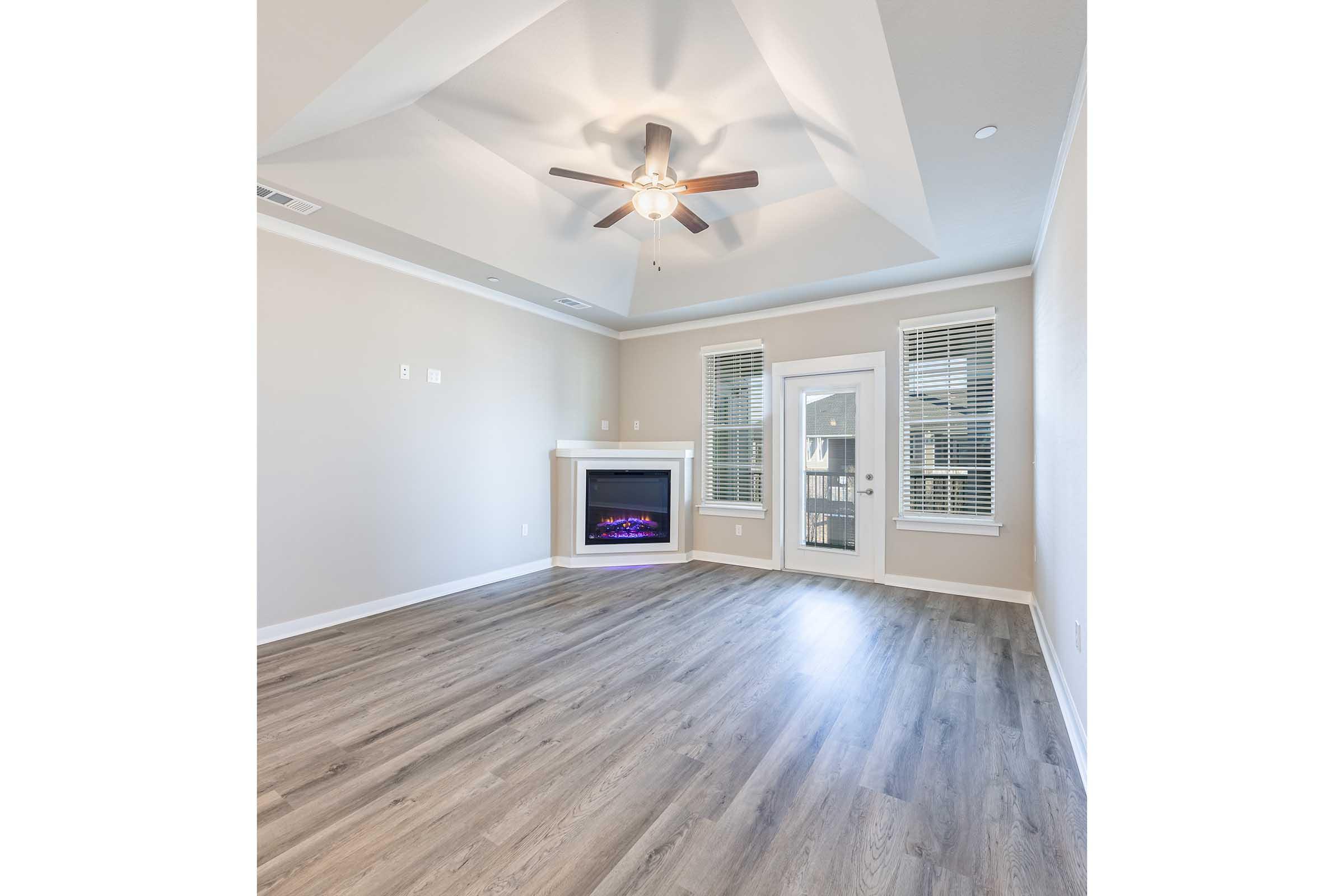 A spacious, well-lit living room featuring a coffered ceiling, ceiling fan, modern electric fireplace with a colorful display, large windows with blinds, and light wood flooring. The walls are painted a neutral color, enhancing the airy feel of the room.
