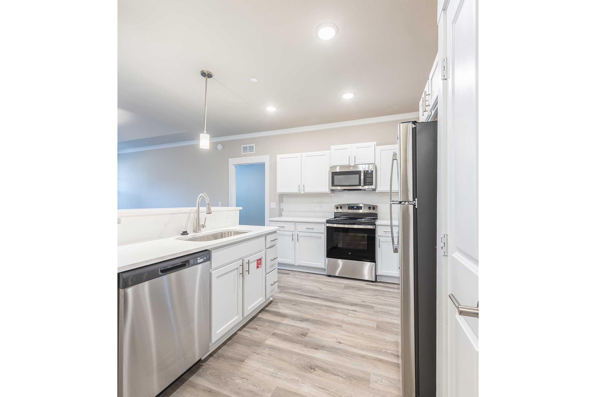 Modern kitchen featuring white cabinetry, stainless steel appliances including a refrigerator, oven, and microwave. A dishwasher is visible, along with a single-basin sink and a contemporary light fixture. The flooring is a light wood finish, enhancing the bright and open atmosphere.