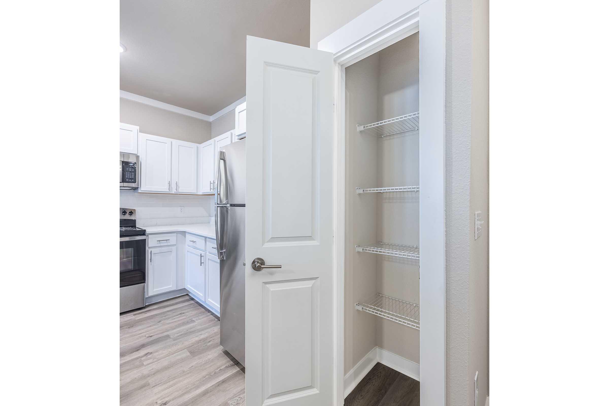 A partially open pantry door revealing empty shelves, with a bright kitchen visible in the background featuring white cabinets, a stainless steel refrigerator, and gray flooring. The kitchen is well-lit and modern, emphasizing a clean and organized space.