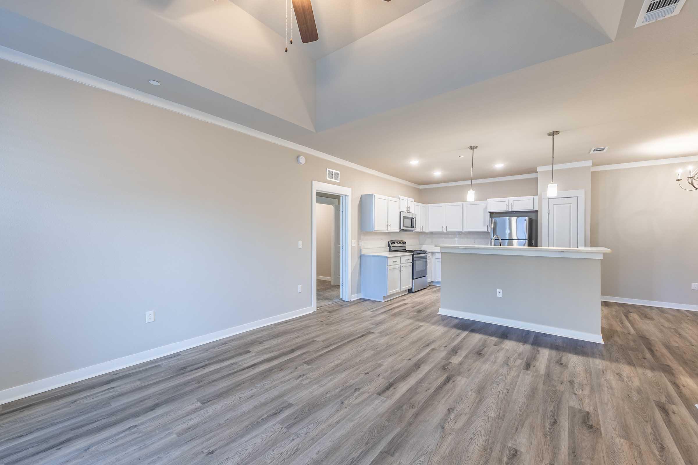 A spacious interior of an apartment with light-colored walls and wooden flooring. It features a modern kitchen with a white countertop, stainless steel appliances, and an open layout leading into a living area. High ceilings with a ceiling fan add to the airy feel. Natural light brightens the space.