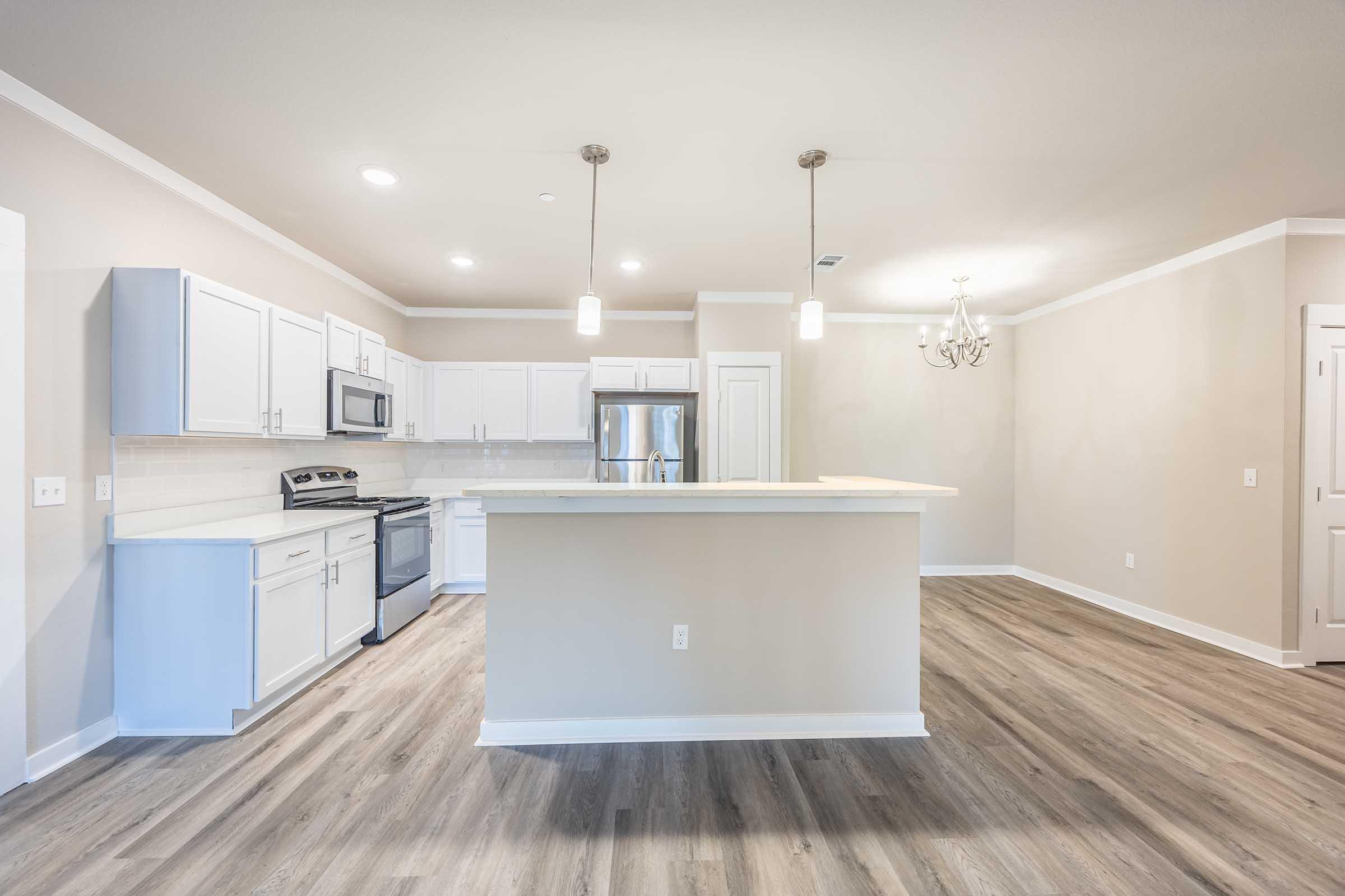 A modern, open-concept kitchen and dining area featuring white cabinets, stainless steel appliances, and a large island with bar seating. The space has light-colored walls, a chandelier, and smooth wooden flooring, creating a bright and inviting atmosphere.