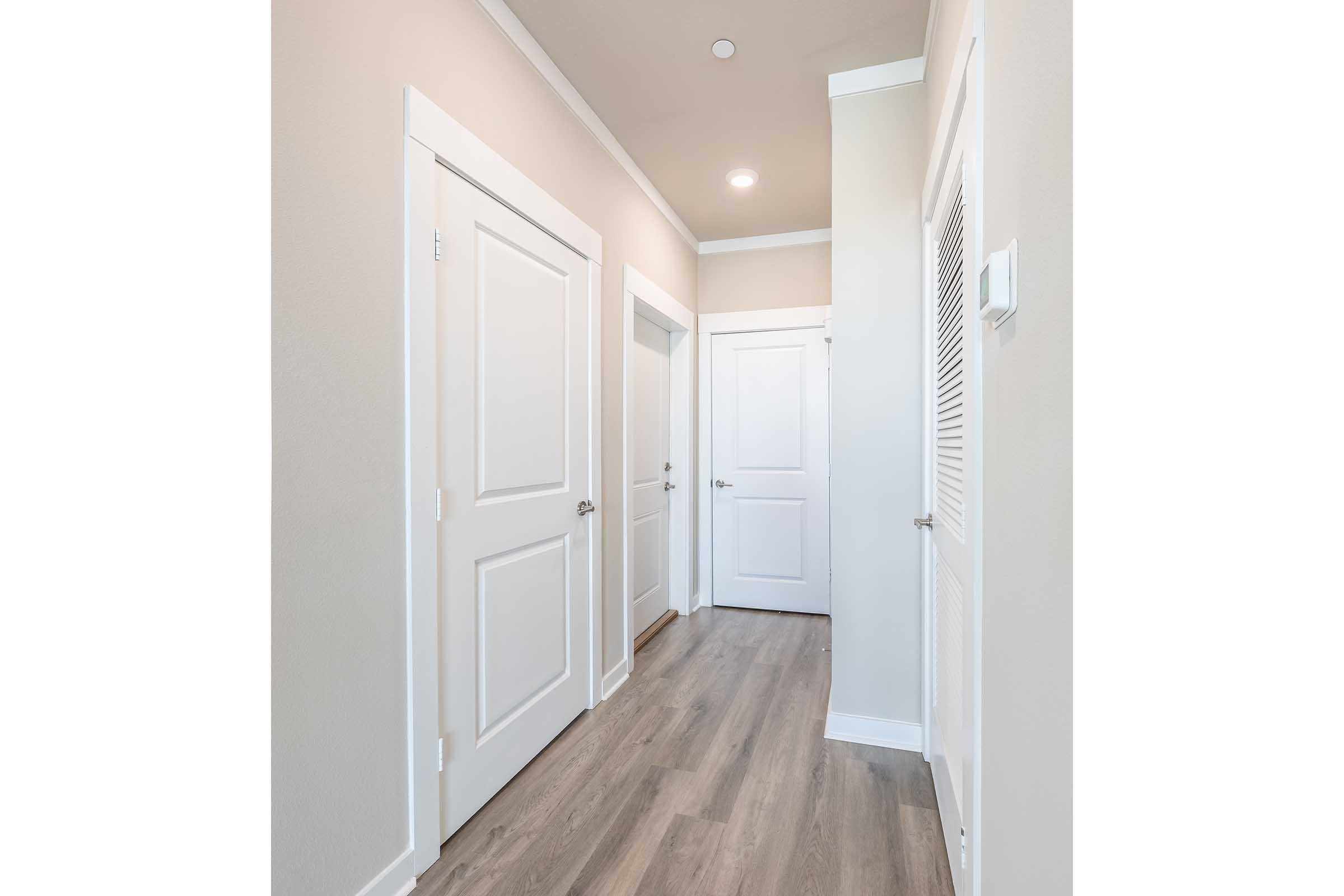A well-lit hallway featuring light-colored walls and wooden laminate flooring. There are three closed doors on the left and right sides, with a vent on the right wall. The ceiling is white with recessed lighting, creating a clean and modern atmosphere.