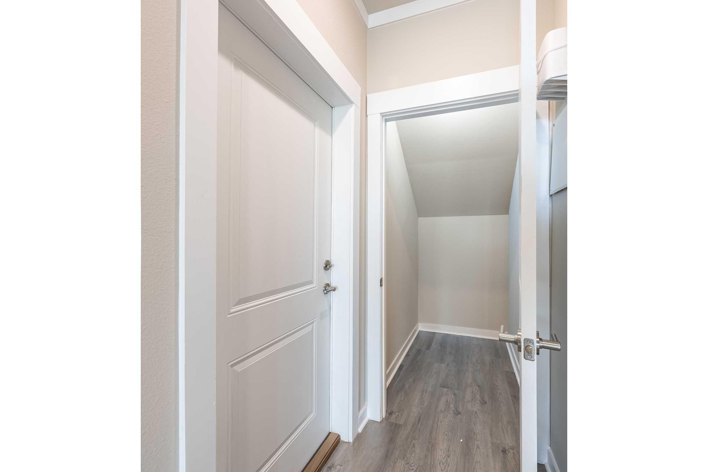 A narrow hallway featuring a closed white door on the left and an open doorway leading to a small storage area on the right. The walls are painted light beige, and the floor is finished with light gray wood-like flooring. The ceiling has a simple design with white trim.