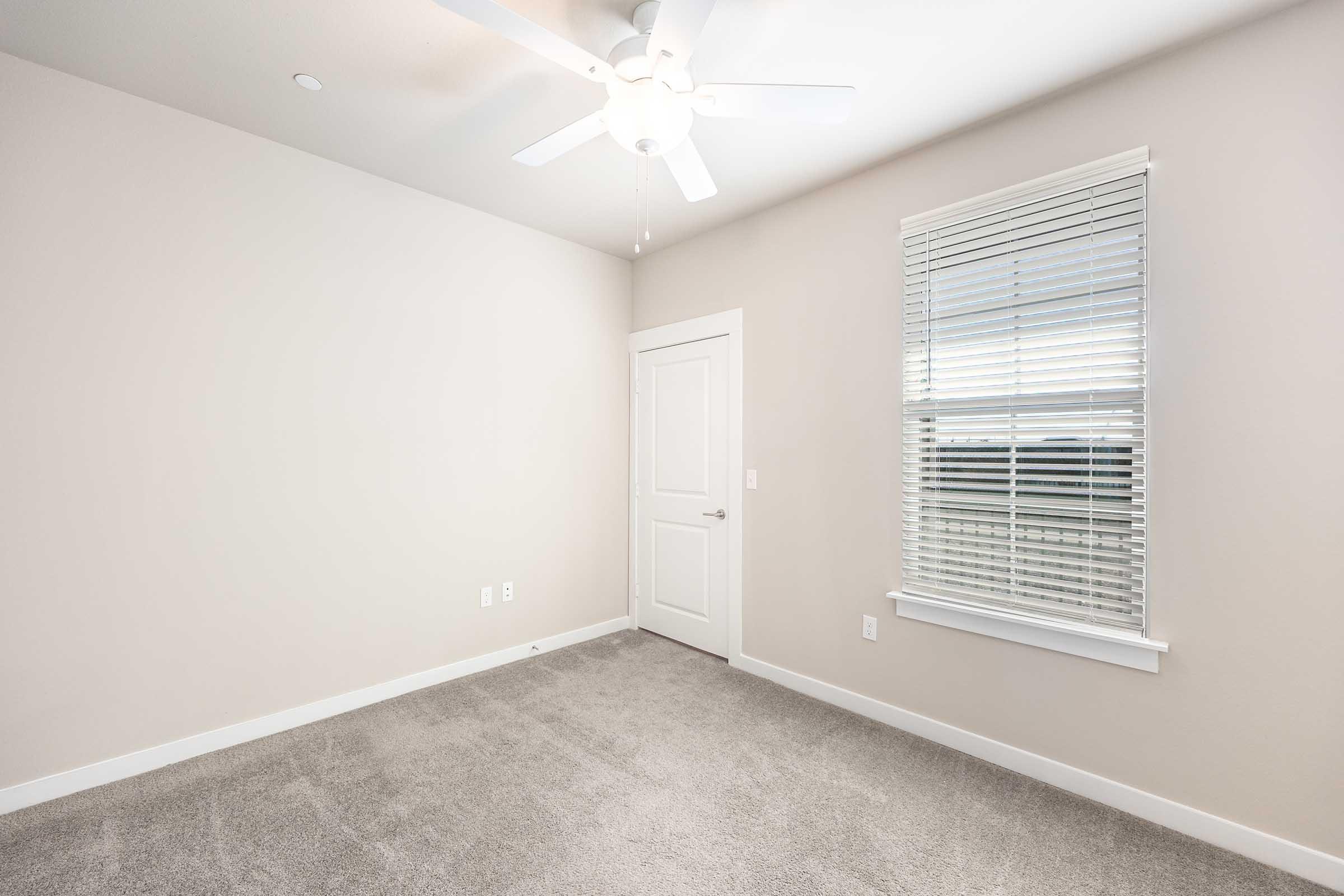 A small, empty room with light gray carpet, a ceiling fan, and a window with blinds. The walls are painted a light color, and there is a closed white door. Natural light is coming in through the window, creating a bright and airy atmosphere.