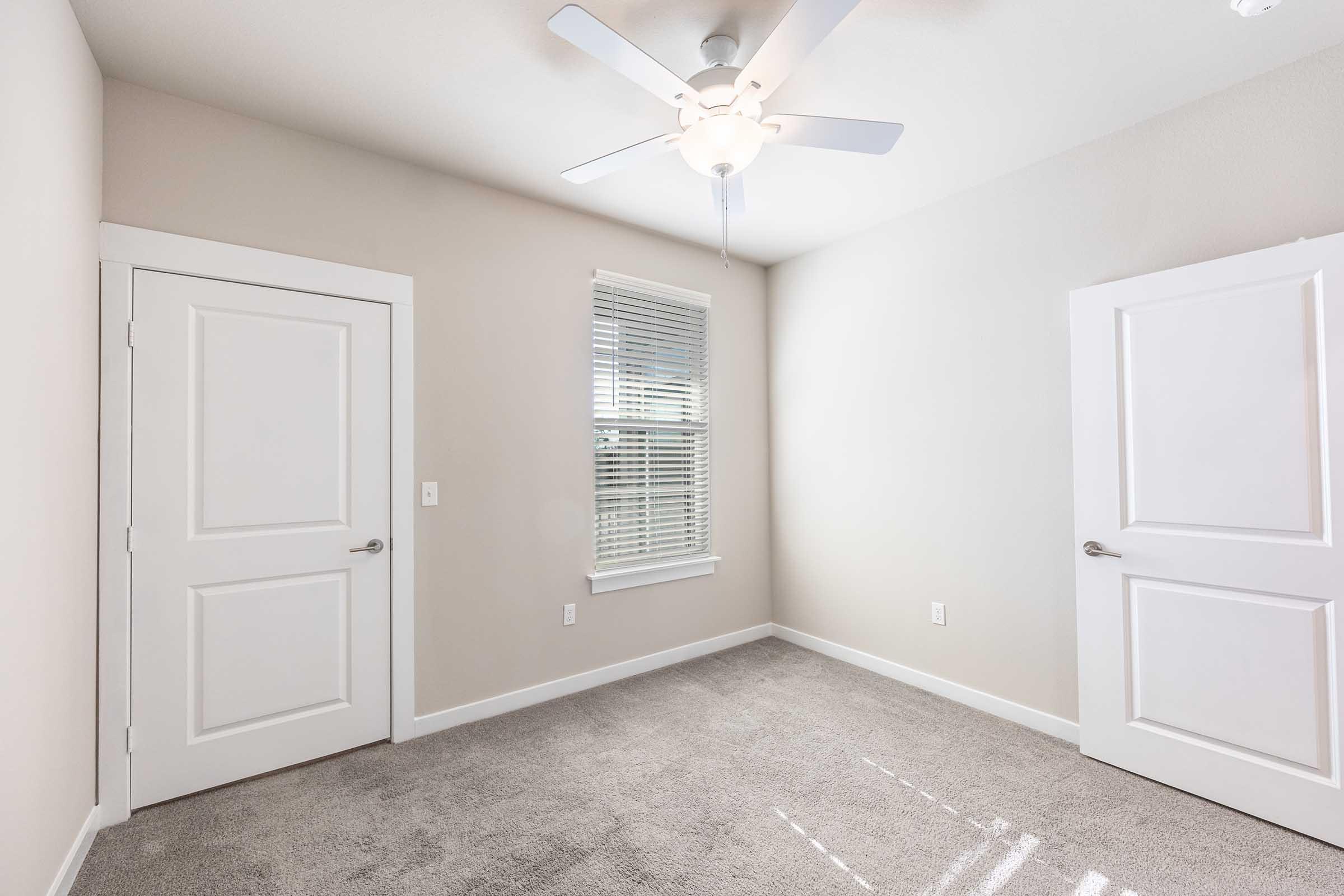 Empty room with light gray carpet, a ceiling fan, and two white doors. One door leads to the outside, while the other is a closet. A window with blinds allows natural light into the space, which features neutral beige walls. Ideal for various uses, such as a bedroom or office.