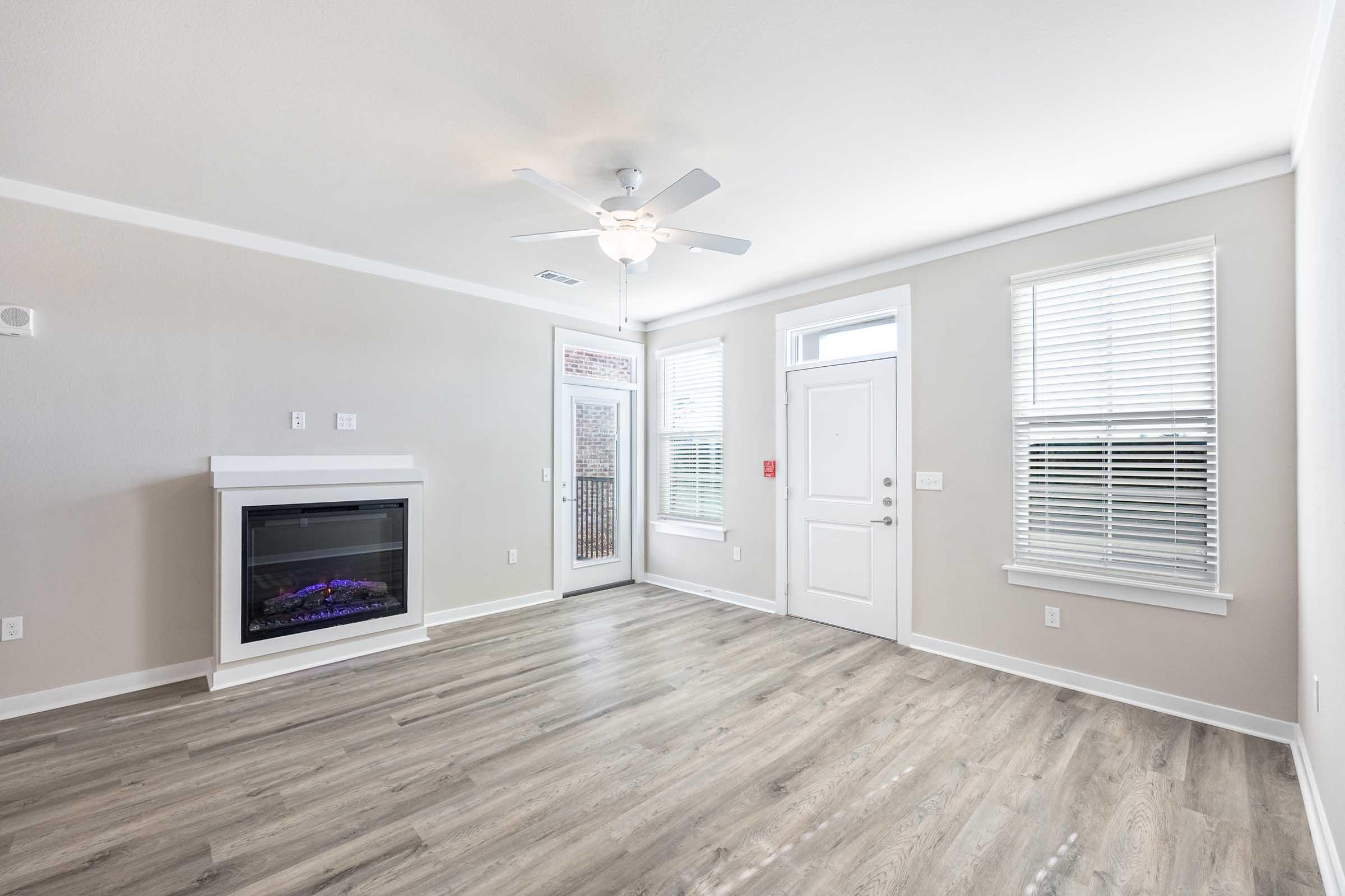 Spacious living room with light-colored walls, a ceiling fan, and a modern electric fireplace. Large windows allow natural light, and there's a front door with a screen. The floor features a wood-like laminate finish, creating a warm and inviting atmosphere.