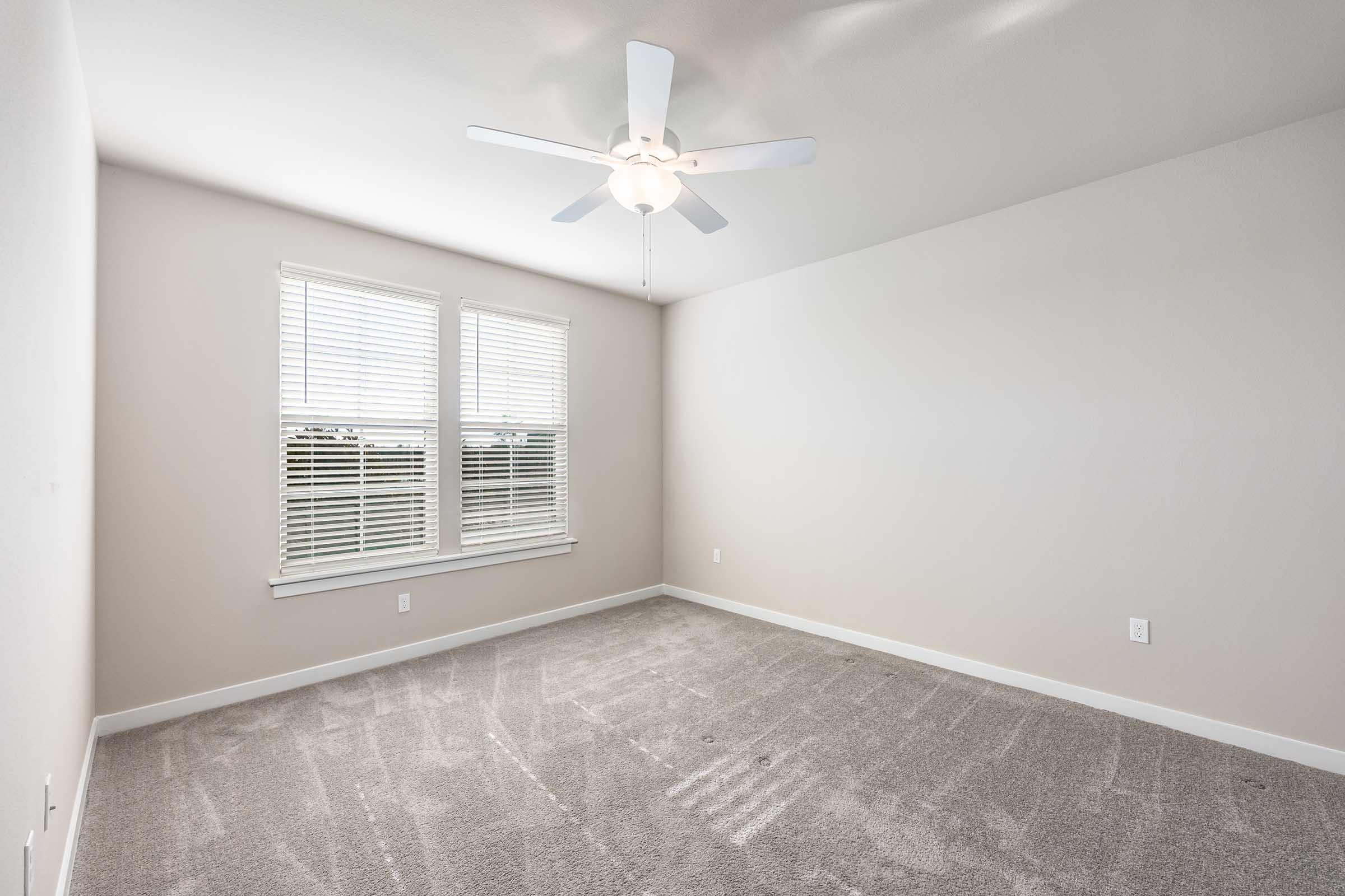 A bright, empty room with light gray walls and a light-colored carpet. There are two windows with white blinds allowing natural light in, and a ceiling fan is mounted in the center of the room. The space is unfurnished and has a clean, minimalistic appearance.