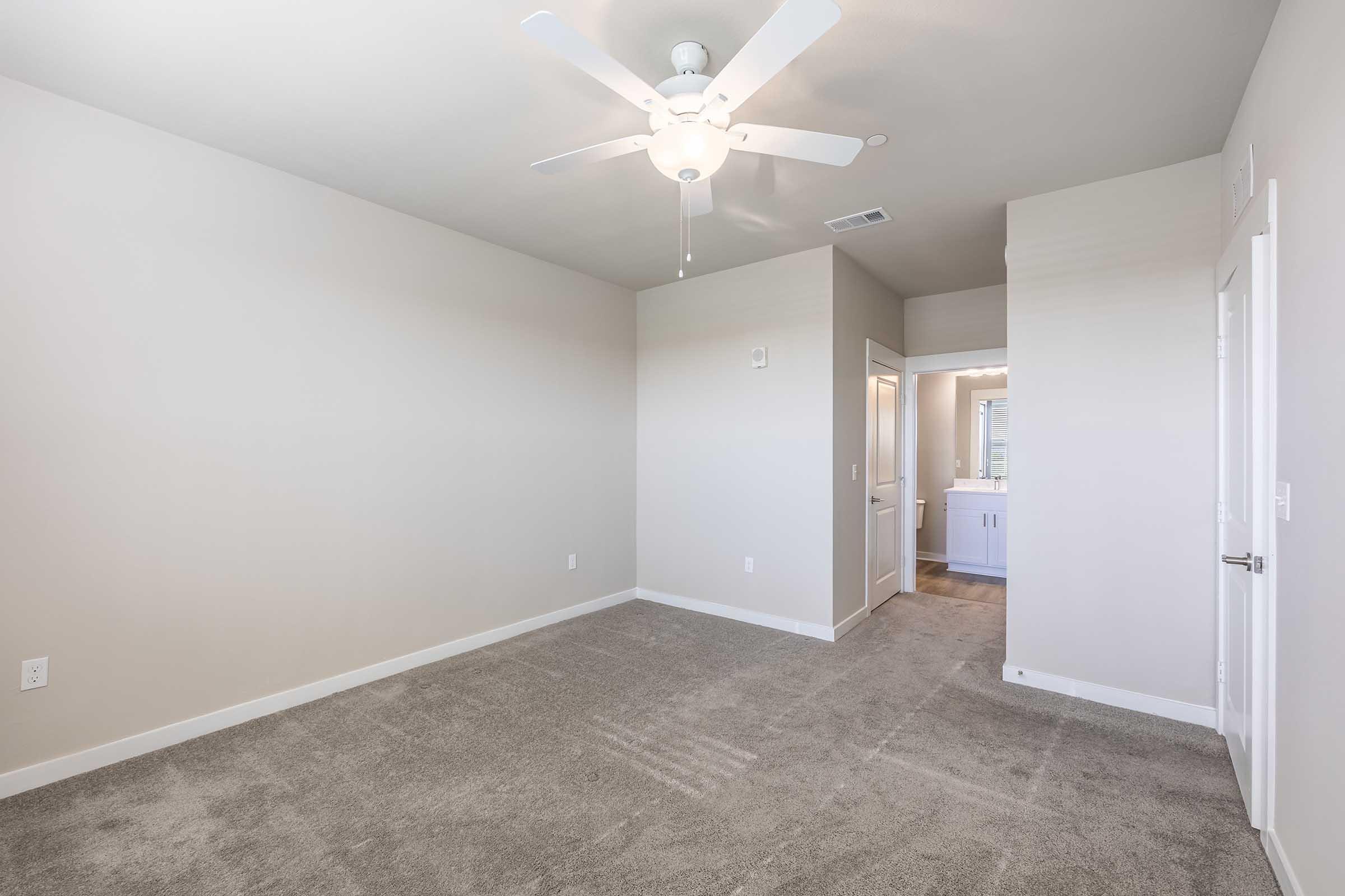 A spacious, empty bedroom featuring light gray carpet and a ceiling fan. The walls are painted in a neutral tone. A doorway leads to a bathroom area, visible at the back, with a glimpse of cabinetry and lighting. Natural light may enter through windows, adding warmth to the room.
