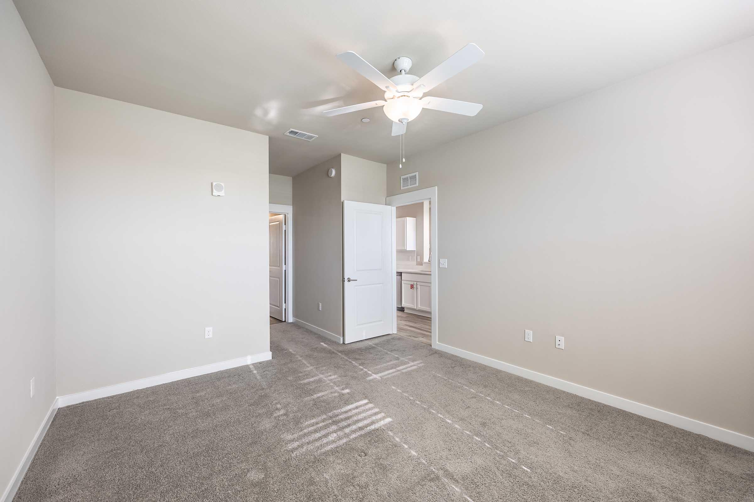 Bright and spacious room with light gray walls and a ceiling fan. The floor is covered in light-colored carpet, and there are two doors leading to adjacent rooms. Natural light streams in from a window, creating a warm and inviting atmosphere.