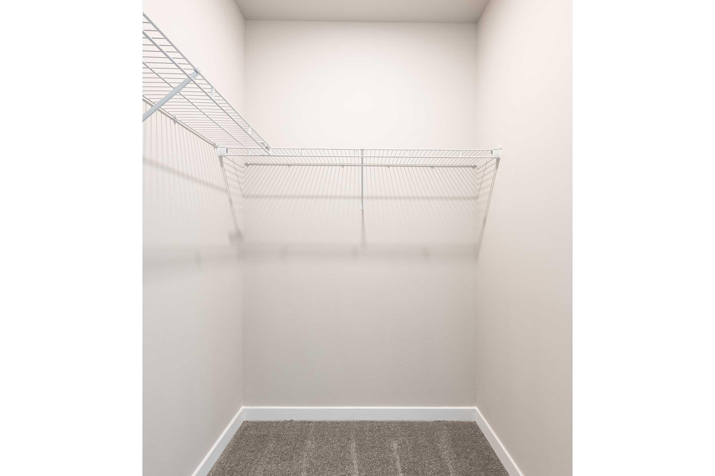 Empty closet with light gray walls and a wire shelving unit installed on the upper left side. The floor is covered with a textured gray carpet, and the space is well-lit, creating an open, minimalist atmosphere.
